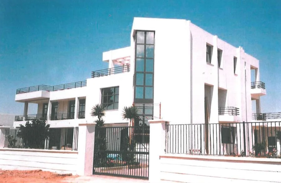 Modern white multi-story residential building with balconies, palm trees, and a black metal fence, under a clear blue sky.