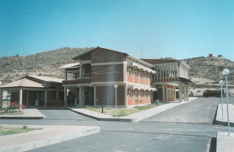 A two-story brick and concrete building with a parking lot in front, surrounded by hilly terrain and a clear blue sky.