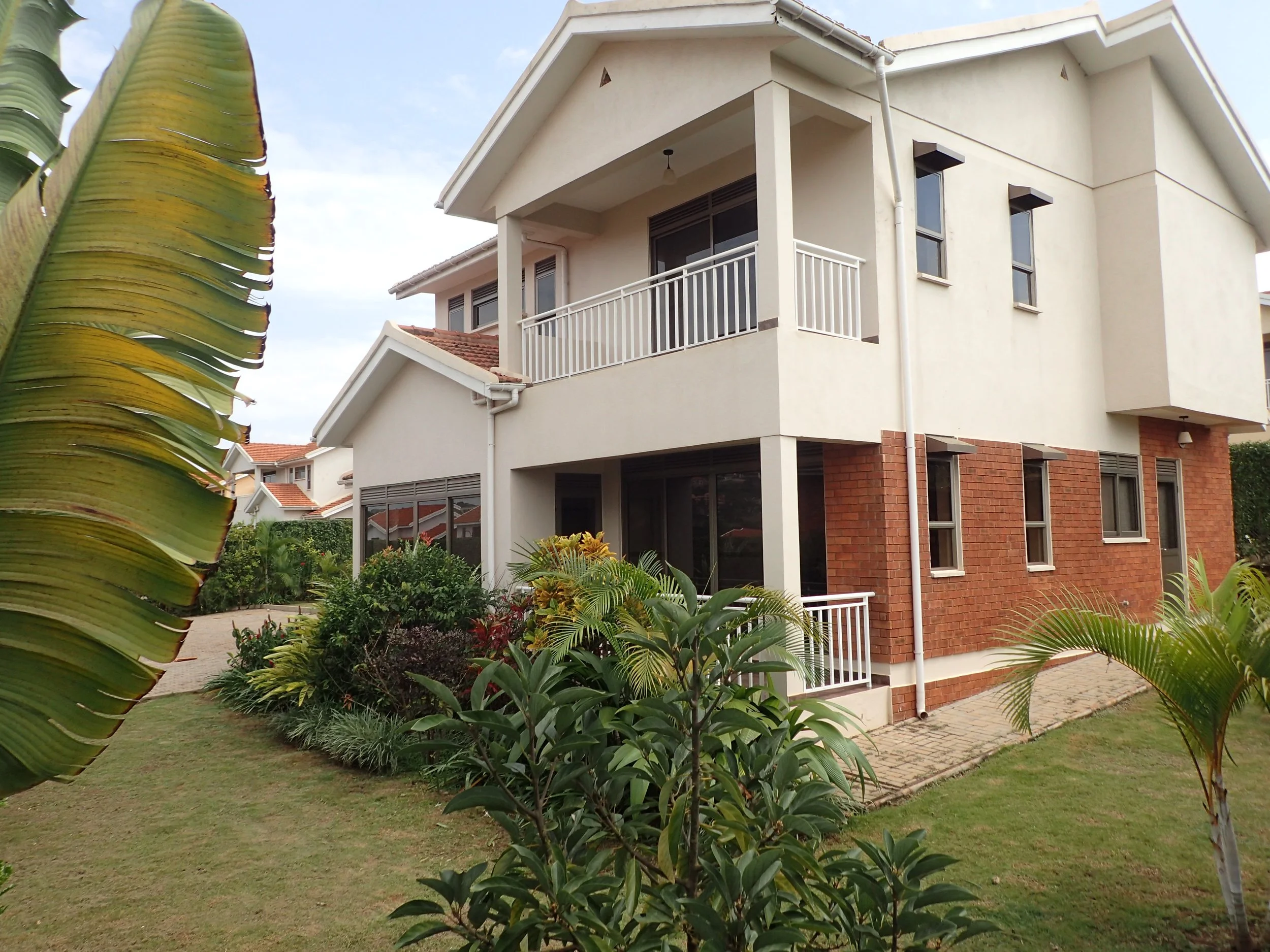 A modern multi-story house with white walls, brick accents, and a balcony with a white railing. The house is surrounded by lush greenery, including various plants, trees, and a grassy yard. The sky is partly cloudy.