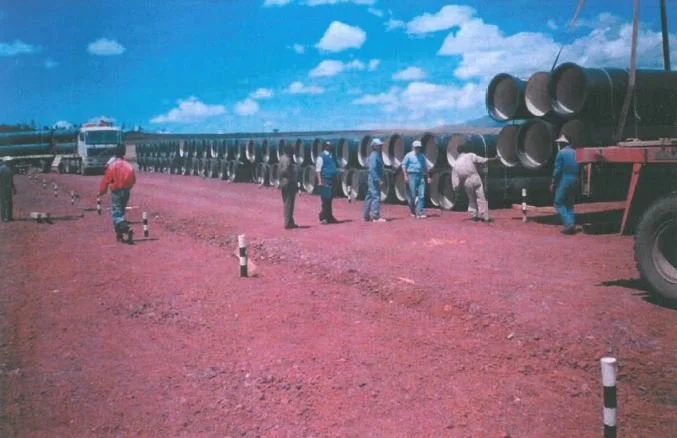 People standing in line to board a train with large pipes in a rural outdoor setting under a partly cloudy sky.