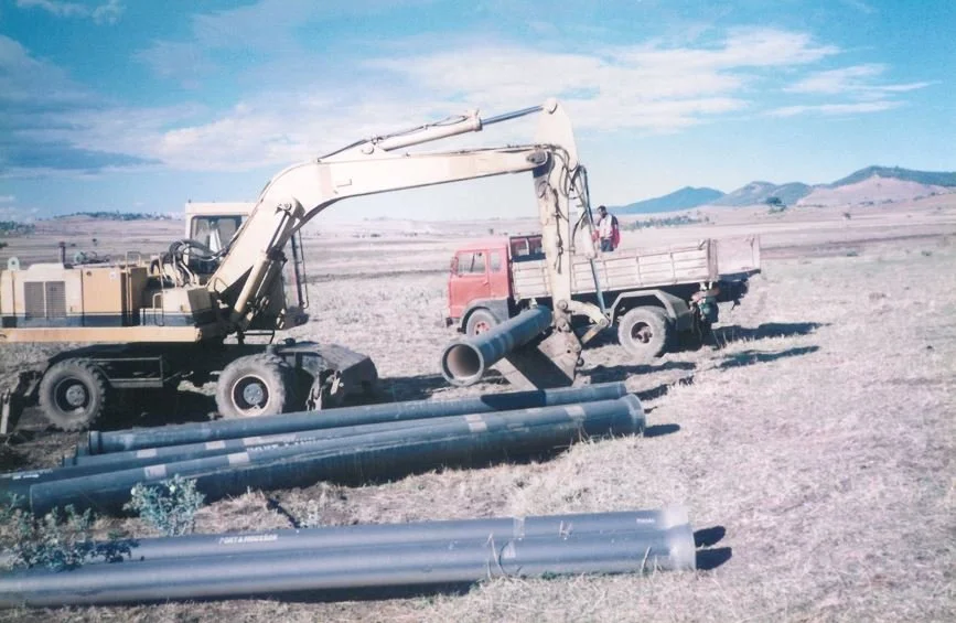 Construction machinery, including an excavator and a flatbed truck, working on installation of large pipes in an open field with mountains in the background.