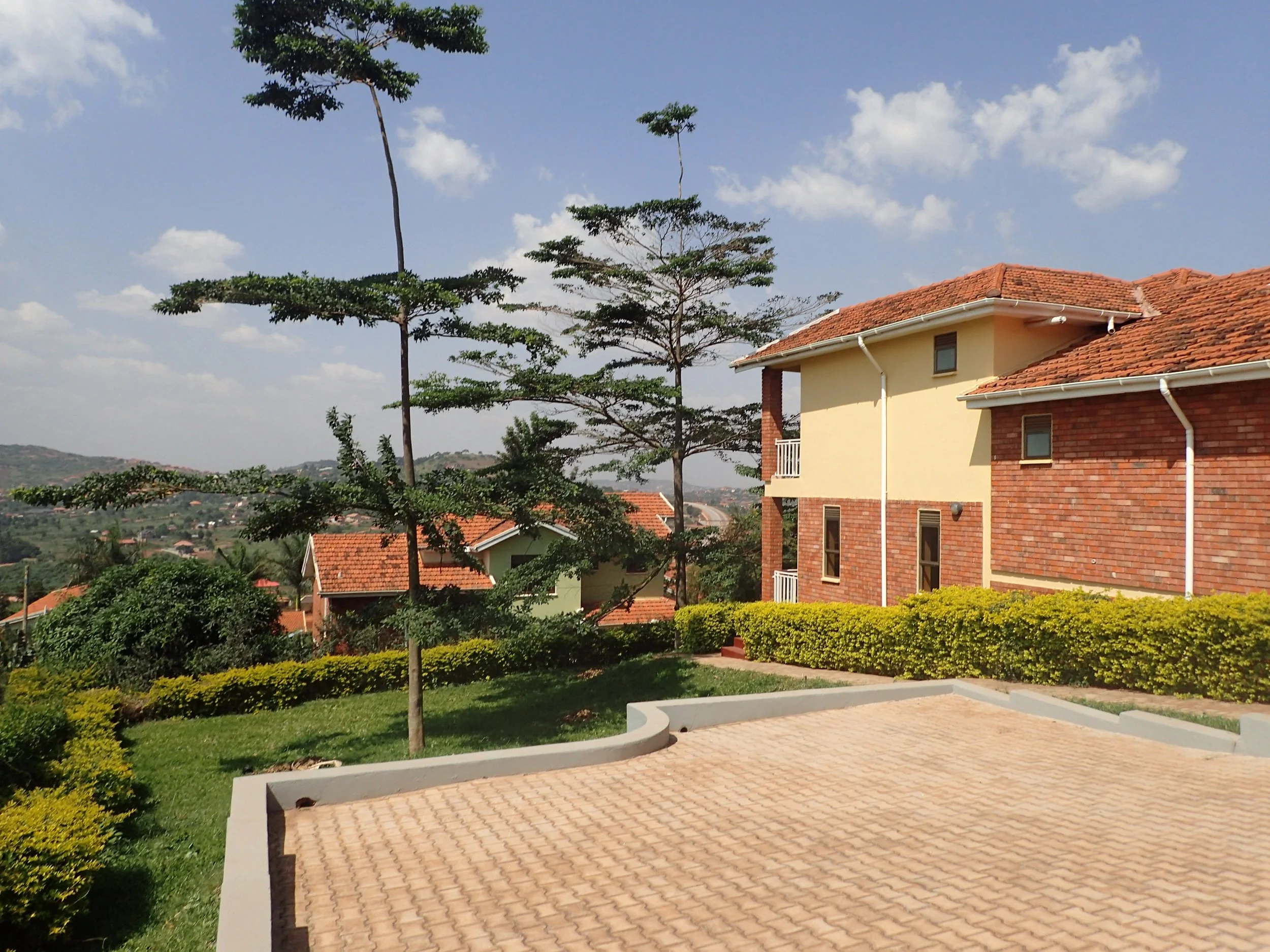A residential backyard with a paved patio area, lush green grass, decorative bushes, a young tree, and a house with a red tiled roof and yellow and brick walls under a blue sky with scattered clouds.