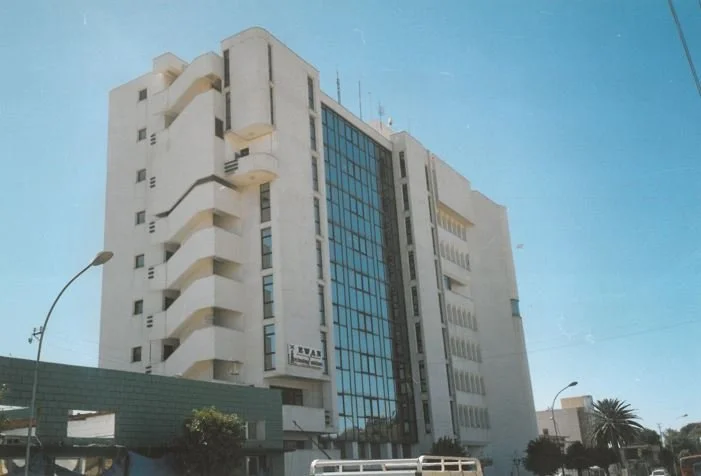A tall white building with curved balconies on its left side and a glass facade on its center, surrounded by street lamps and trees, under a clear blue sky.