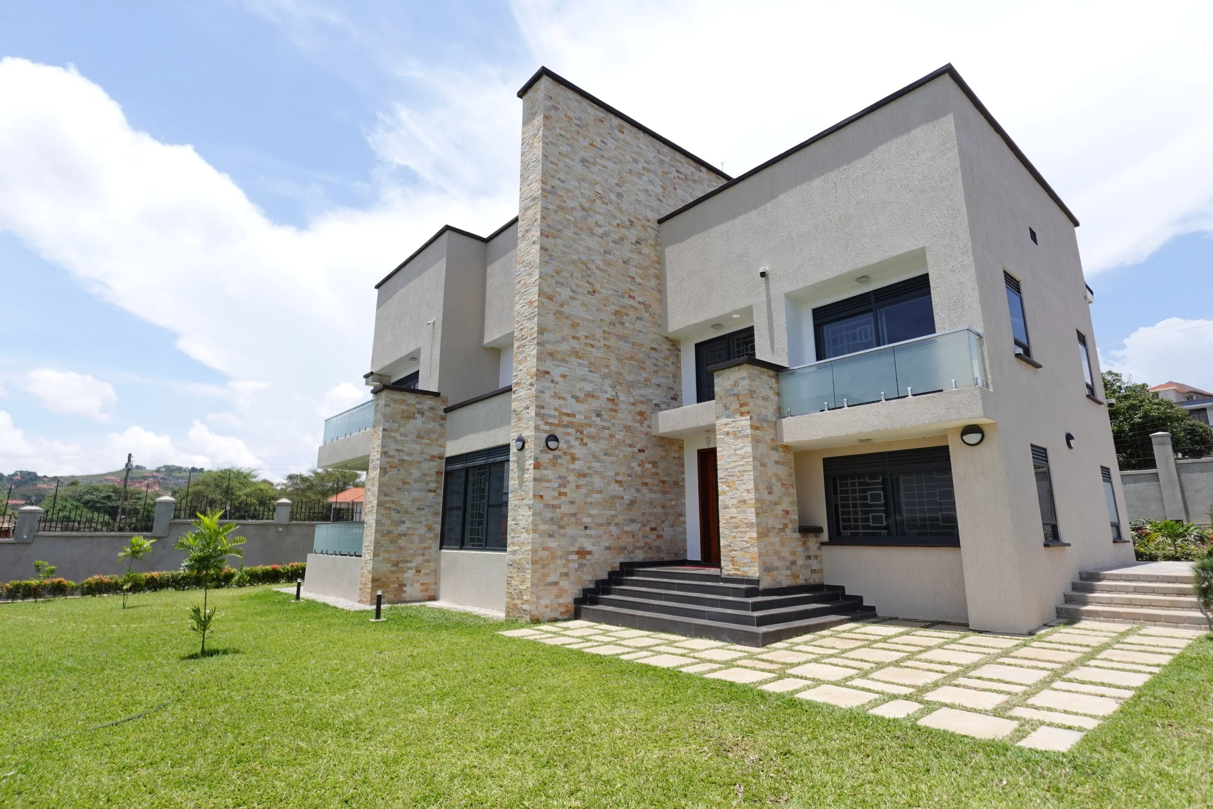 Modern two-story house with a white exterior, stone accents, glass balcony, and black window frames, set against a blue sky with scattered clouds.