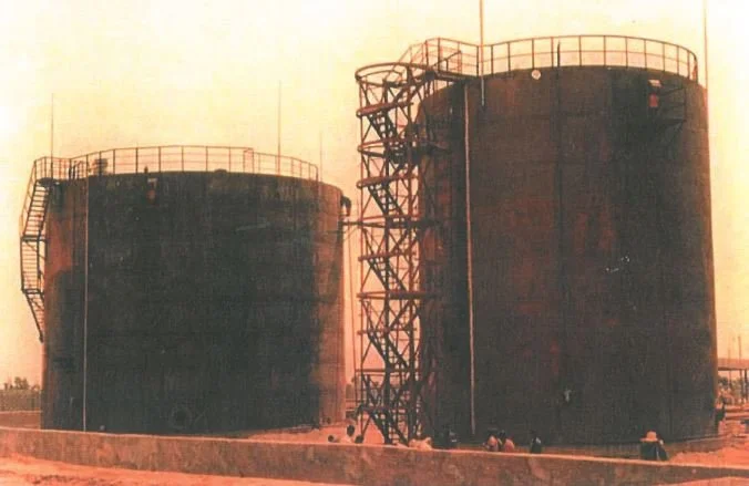 Two large, cylindrical, metal storage tanks with external staircases and railings, set in an industrial area under a hazy sky.