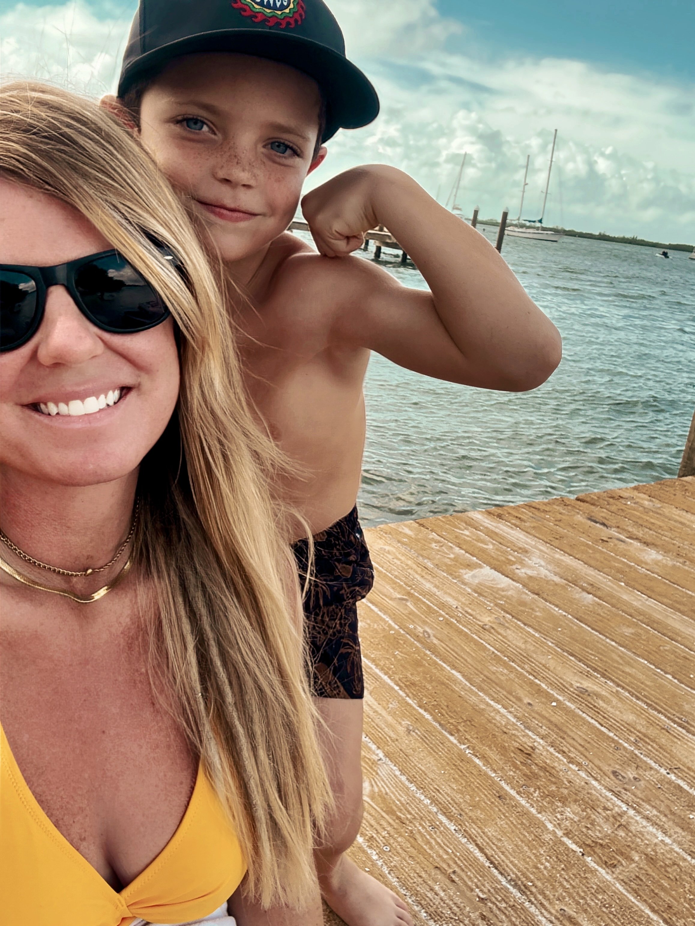 Young boy flexing his bicep and smiling at the camera near water with boats and a dock, woman with blonde hair and sunglasses in the foreground, wooden deck in the bottom right.