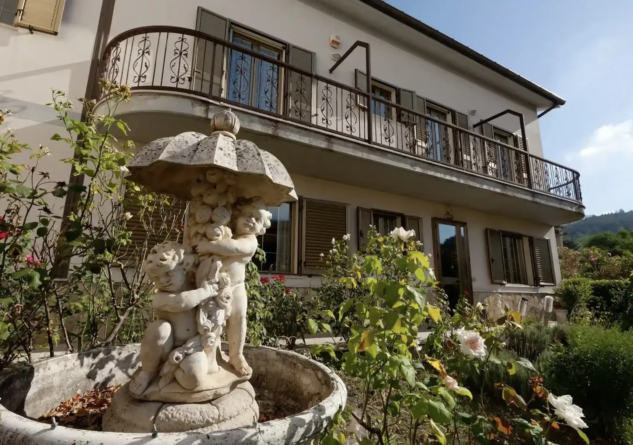 A decorative stone fountain featuring cherub figures in a garden surrounded by blooming flowers, with a multi-story house in the background.