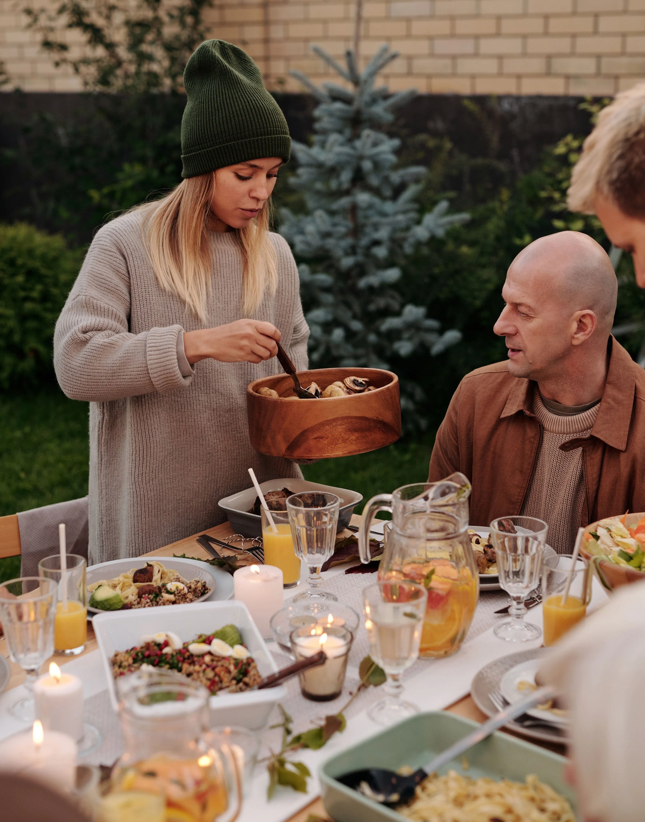A woman in a green beanie and beige sweater serving food at an outdoor dinner table with various dishes, drinks, and candles as background.