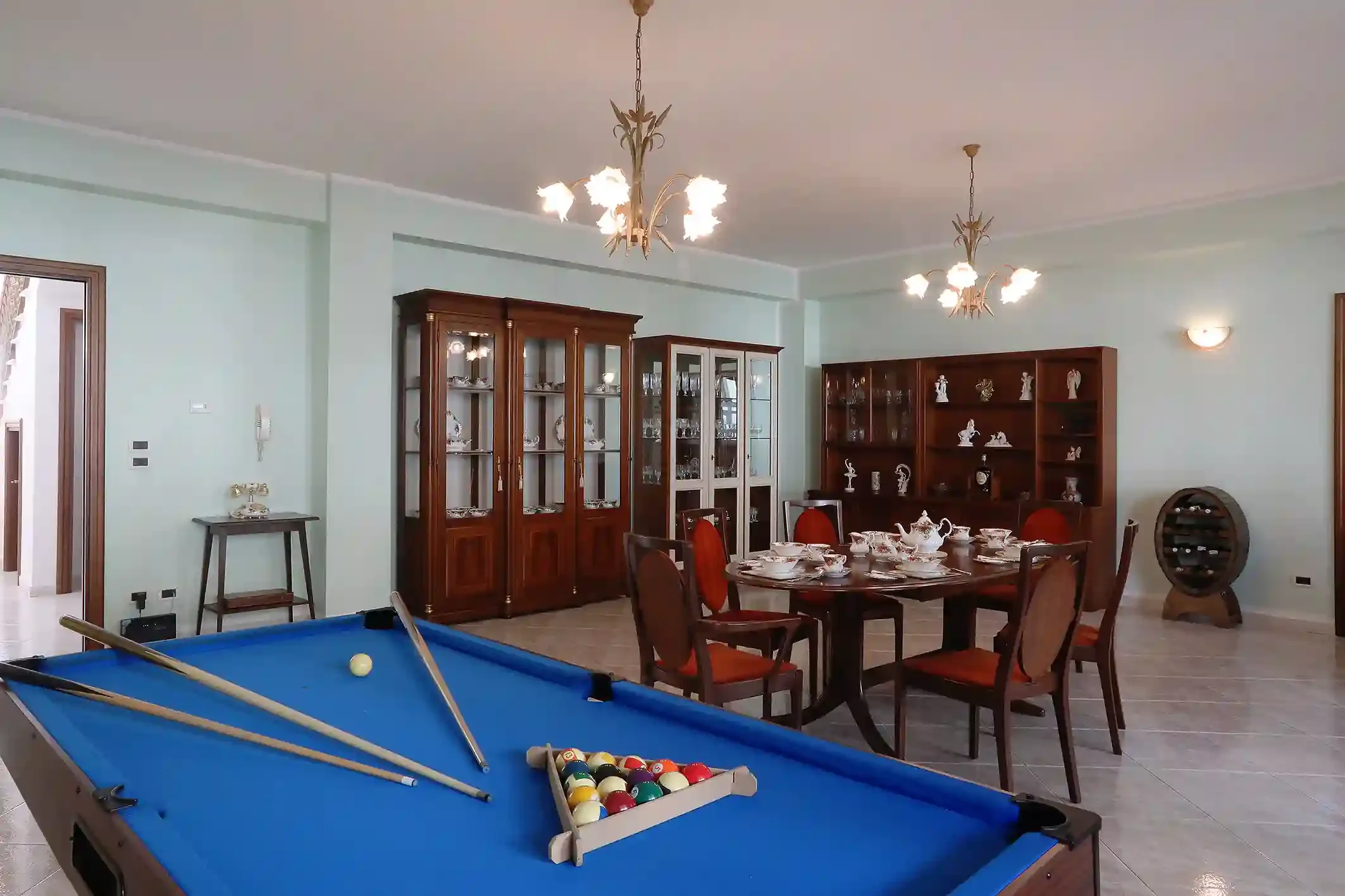 A blue pool table with pool balls arranged, cue sticks laid across, in a room with wooden china cabinets filled with glassware and decorations, a dining table set with teapots and cups, and a chandelier hanging from the ceiling.