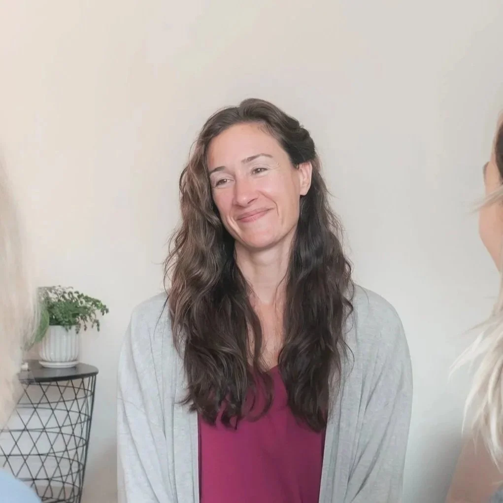 A woman with long, wavy brown hair and a light gray cardigan smiling while talking to two other women in a plain room with a small table and potted plant in the background.