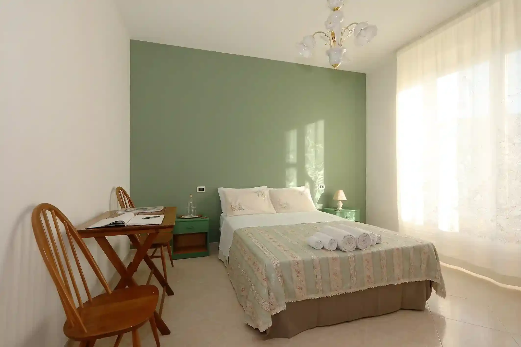 A bedroom with a double bed, three rolled towels on it, a small green nightstand, a glass water bottle, and a table with three wooden chairs. Light curtains cover the window, and there's a chandelier hanging from the ceiling. The room has a green accent wall and white walls, with a beige and light-colored bedding.