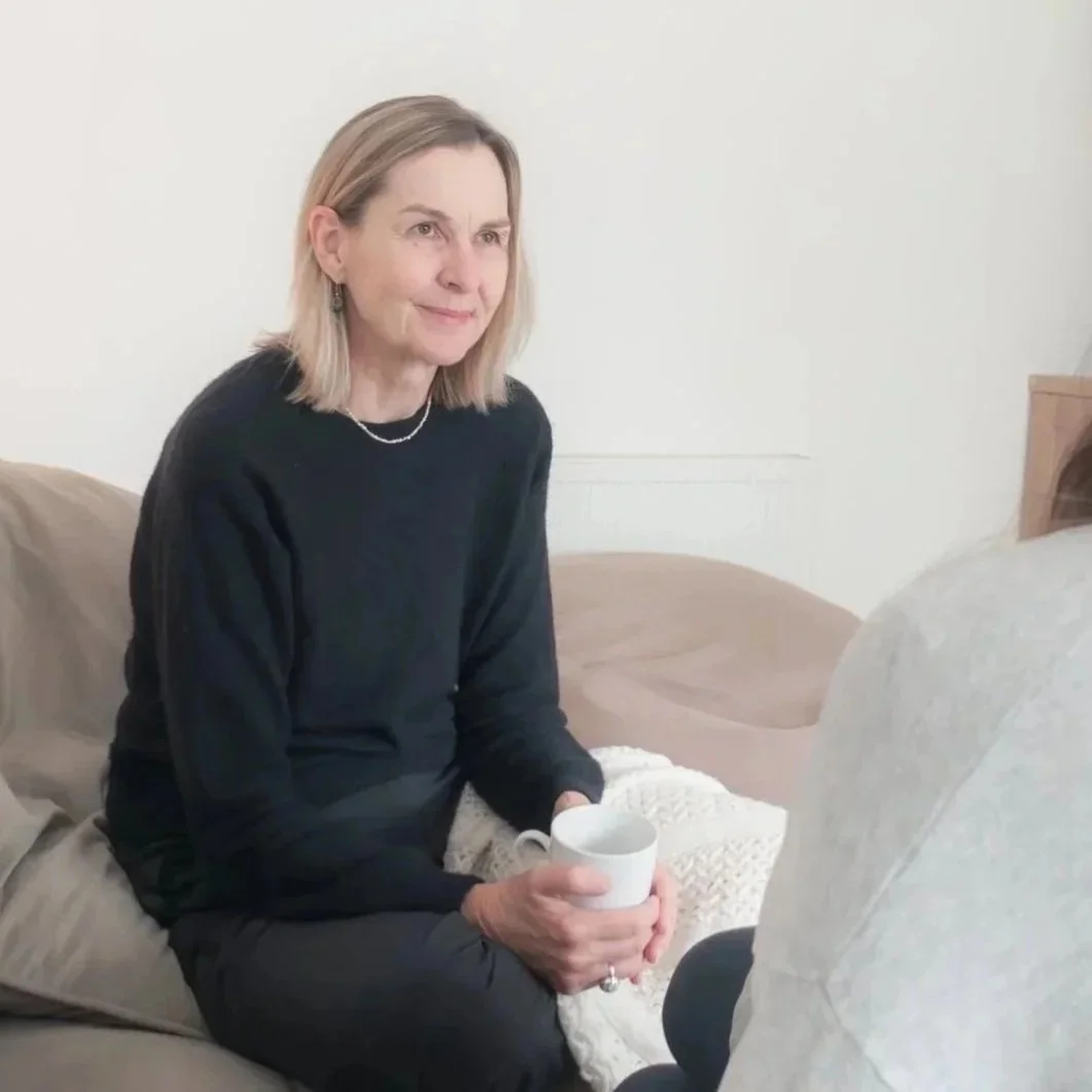 A woman with shoulder-length blonde hair, wearing a black sweater and small earrings, sitting on a beige sofa, holding a white mug with both hands, and looking slightly to her right with a gentle smile.