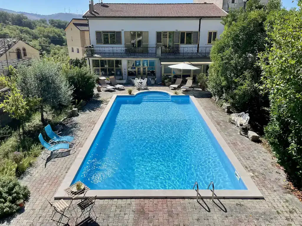 A rectangular outdoor swimming pool with blue water, surrounded by a brick patio and lush green trees and shrubs. There are lounge chairs, a table with an umbrella, and some chairs on the patio, with a two-story house in the background.