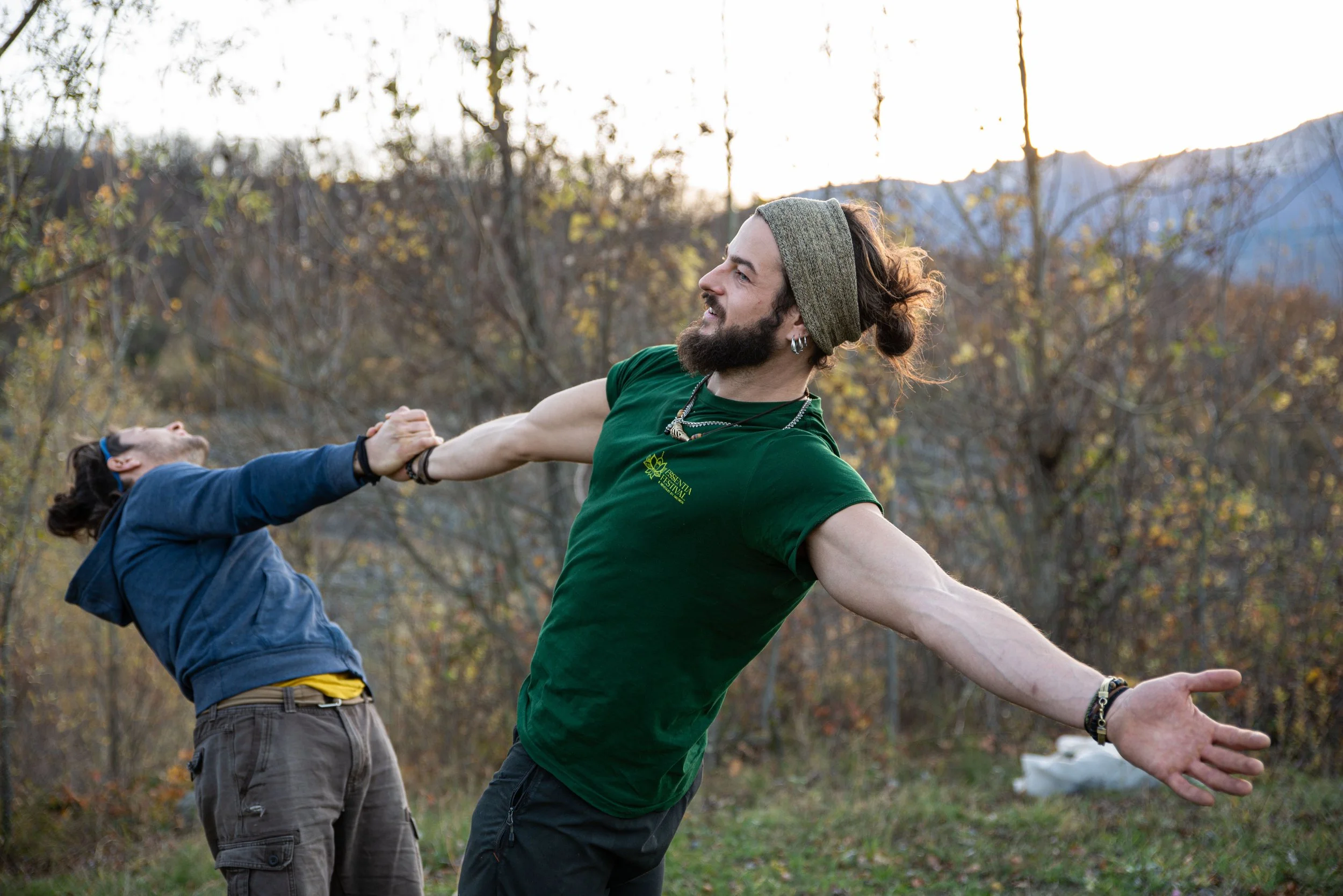 Two men outdoors, one with a beard and long hair wearing a headband and a green shirt, the other with short hair and a blue jacket, holding hands or pulling on each other during daytime.