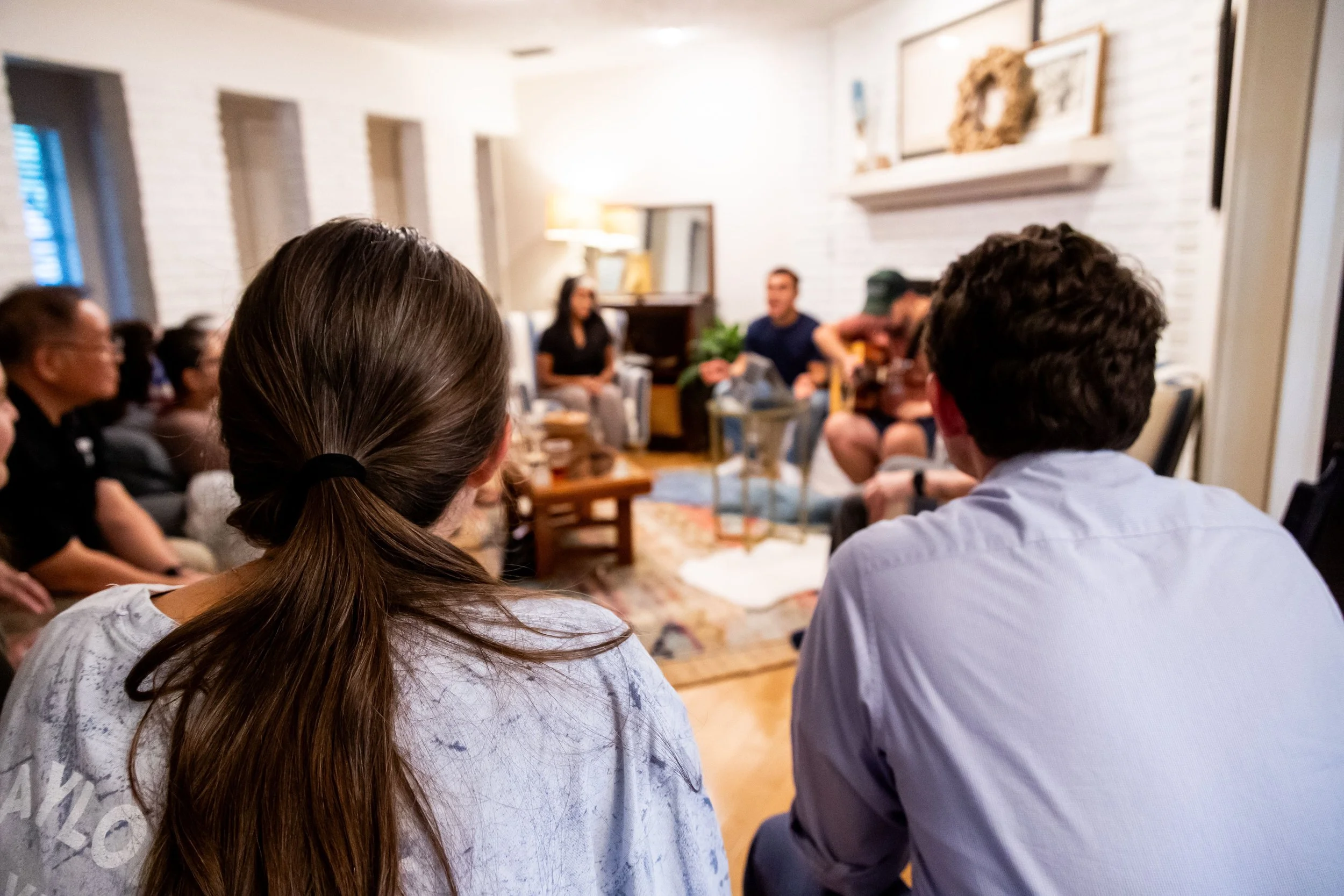 Group of people sitting in a living room, listening to a woman speaking. The room has white brick walls, a fireplace, and framed artwork.