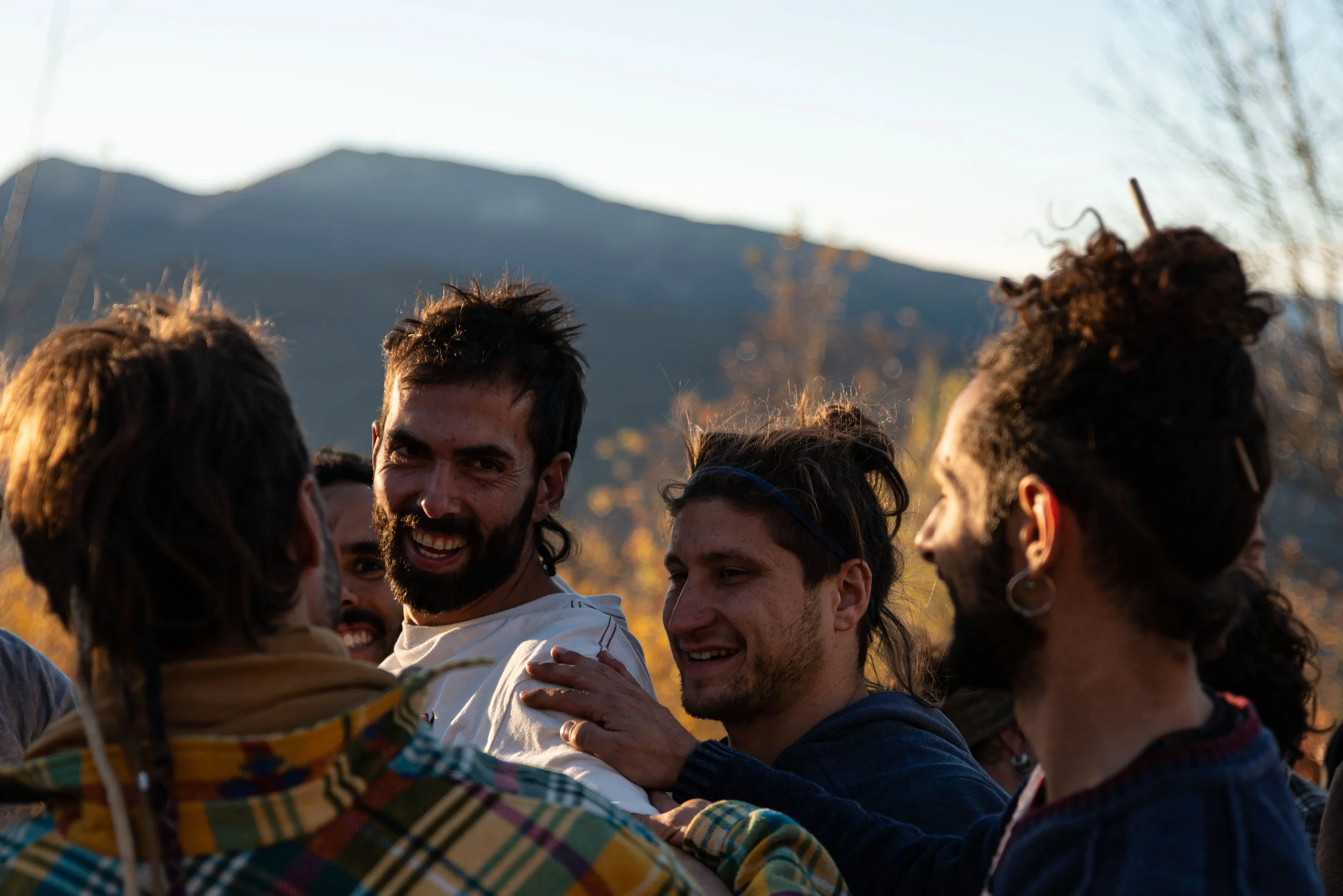 Group of smiling young adults outdoors, embracing each other with arms around shoulders, with mountains and trees in the background during sunset.