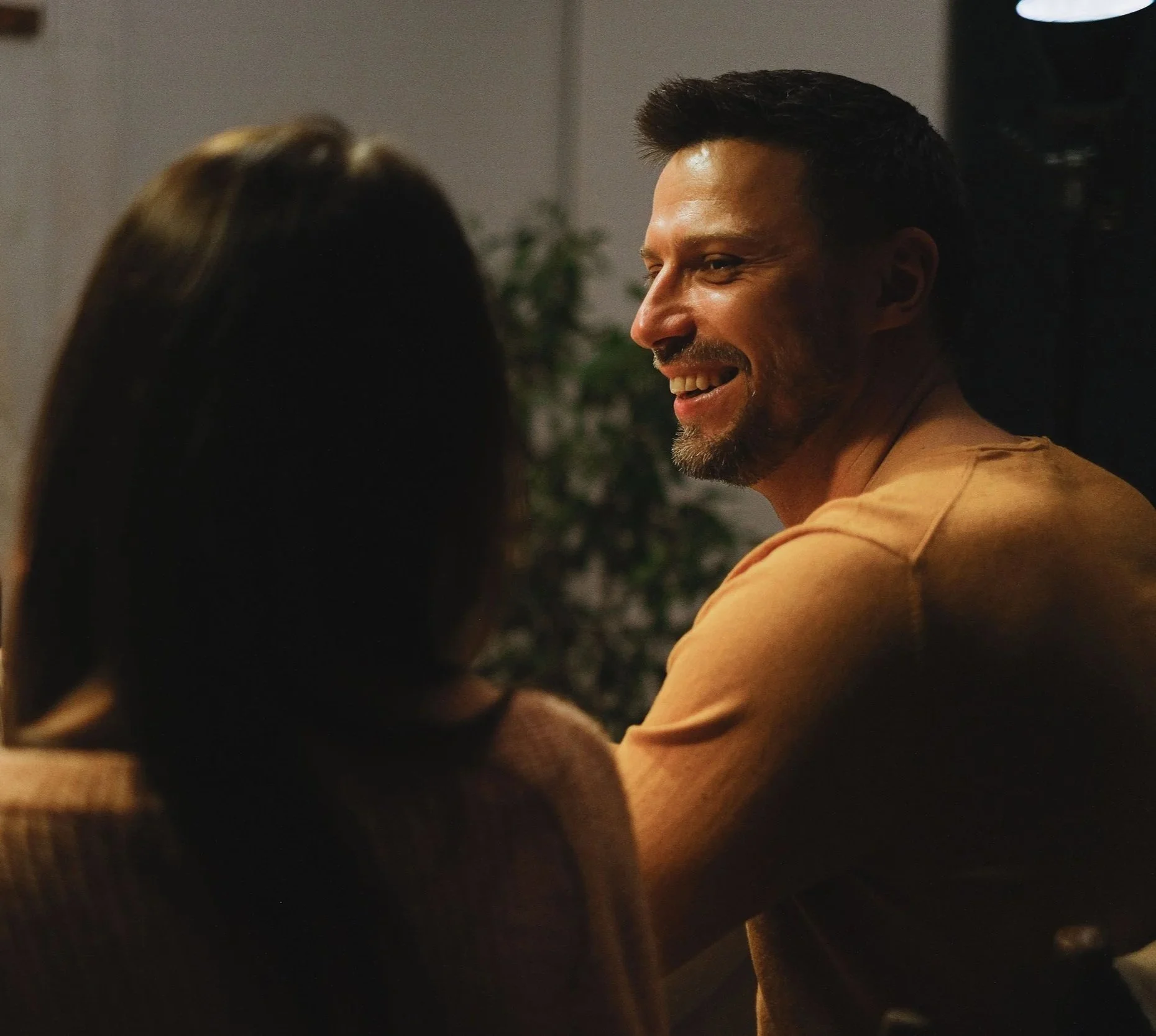 A man with dark hair and a beard smiling, viewed from the side, with a woman with brown hair and a tan top, indoors, with a plant in the background.
