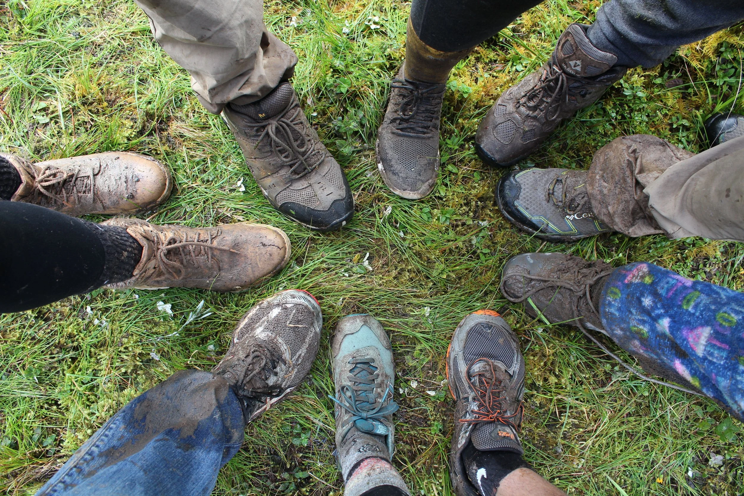 Multiple muddy hiking boots and shoes arranged in a circle on grass.