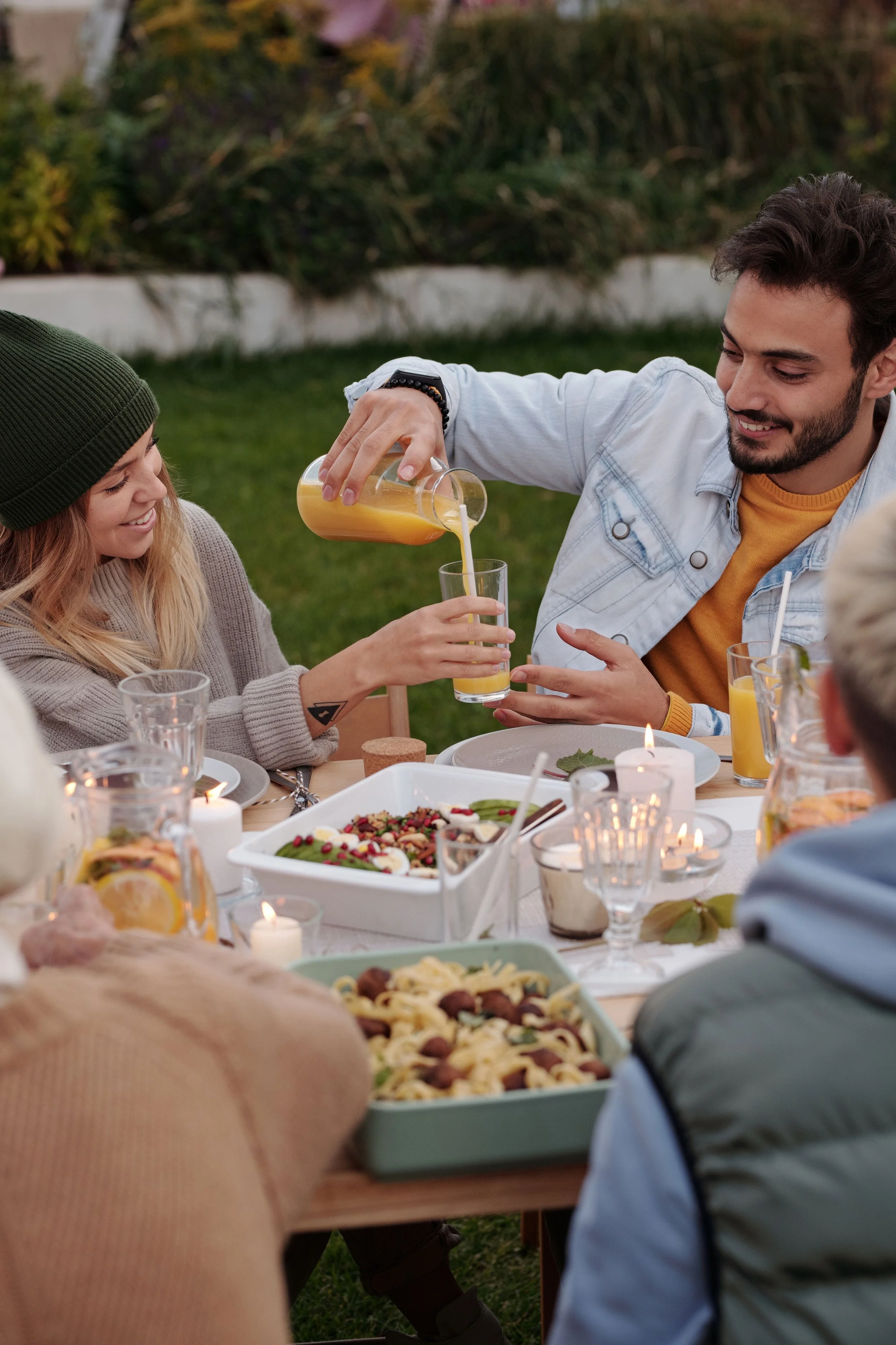 People enjoying an outdoor dinner party, with food and drinks on the table, and a person pouring orange juice into a glass.
