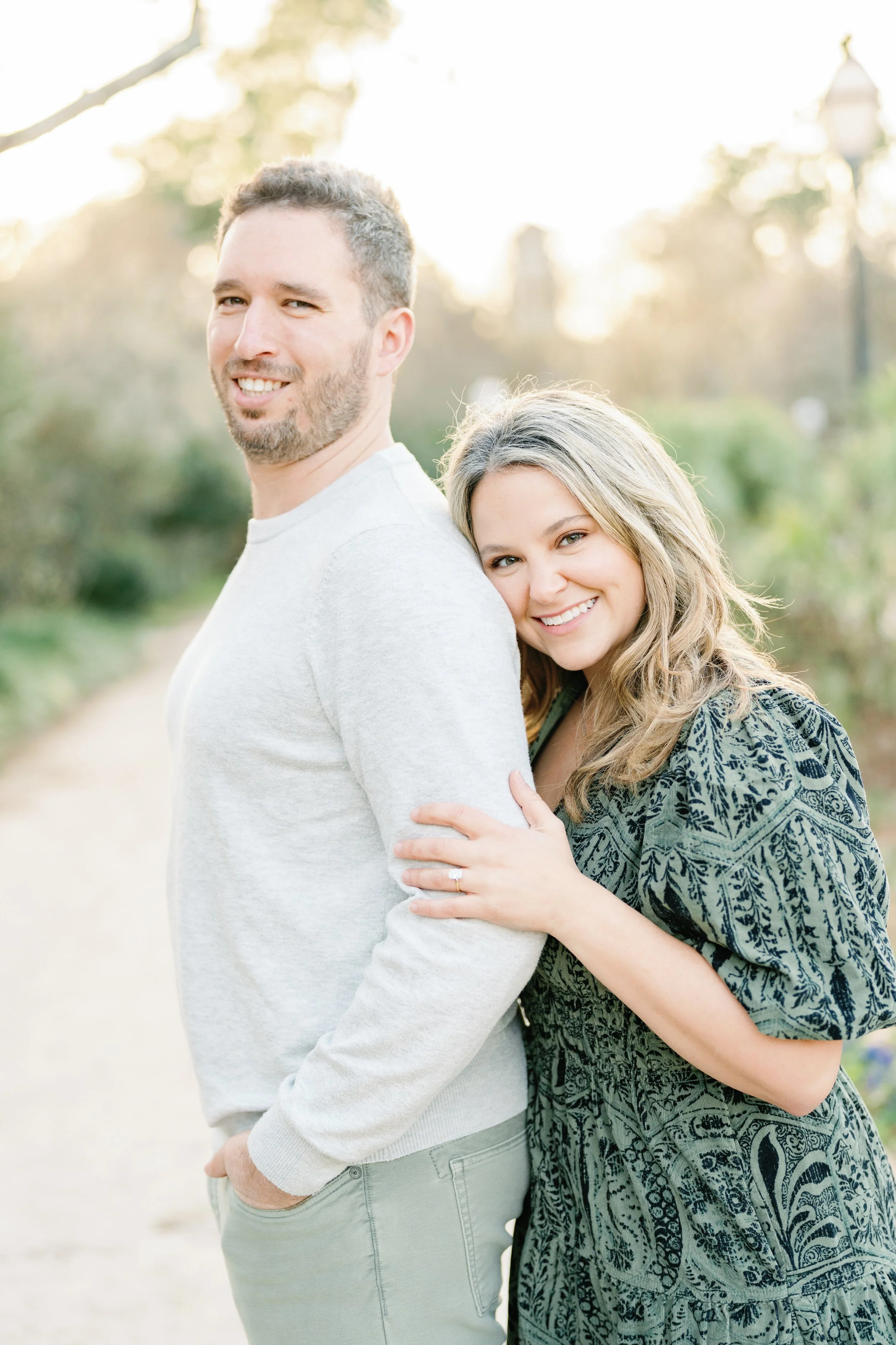A smiling man stands with a woman hugging his arm, outdoors with trees and soft sunlight in the background.