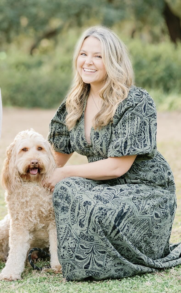 A smiling young woman with long blonde hair kneeling next to a light-colored, curly-haired dog outdoors in a park or garden setting.