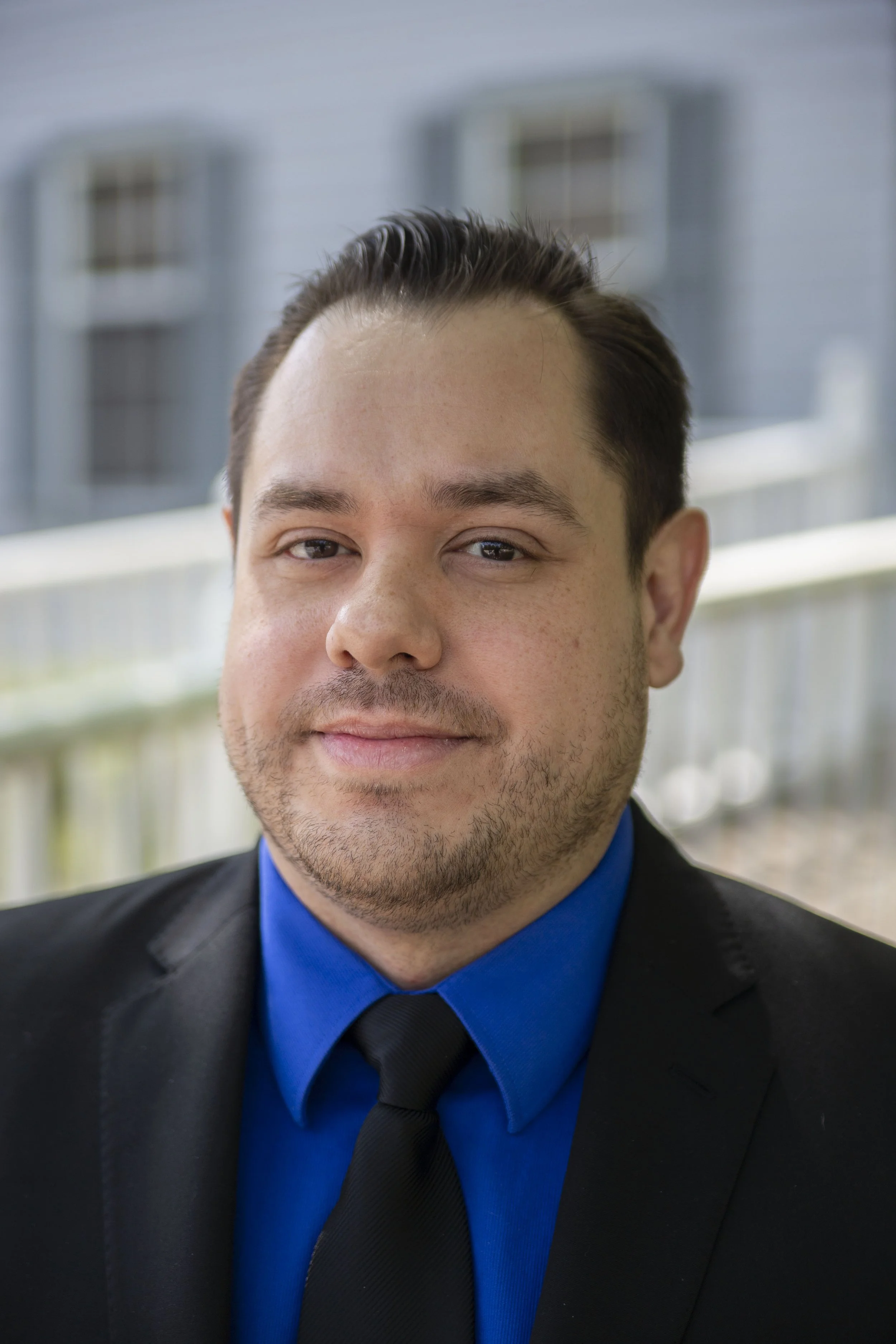 Close-up of a young man with light skin, wearing a black suit, blue shirt, and black tie, smiling outdoors with blurred buildings in the background.