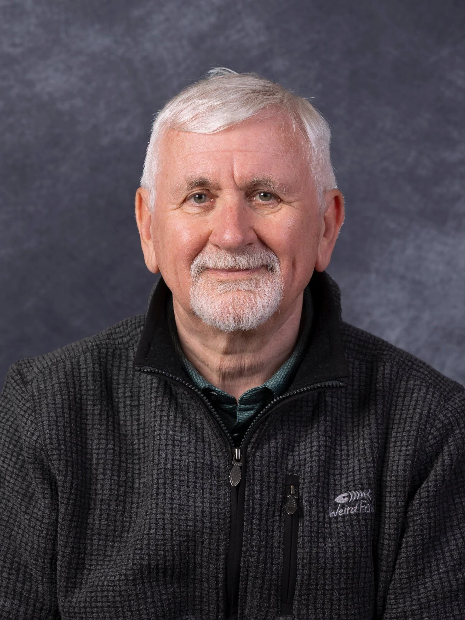 Portrait of an elderly man with white hair, a beard, wearing a black zip-up jacket with a 'Weird Fish' logo, set against a dark background.