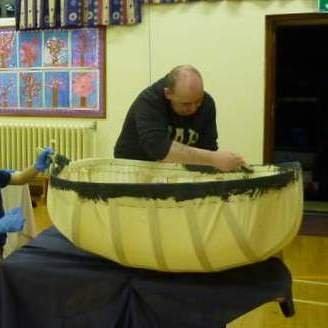 A man working on a large pumpkin that is cut in half, likely carving or preparing it, with a child nearby in a room decorated with a window showing a winter scene.