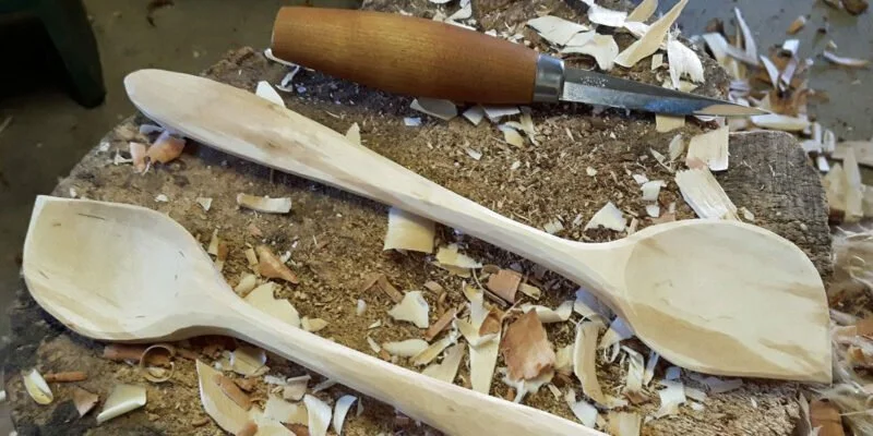 Two wooden spoons and a wood carving tool on a work surface covered with wood shavings.