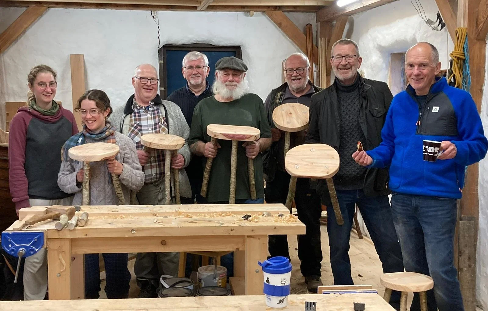 Group of eight people standing inside a woodworking shop, holding completed wooden stools, with a workbench and wood shavings in front, smiling and posing for the photo.