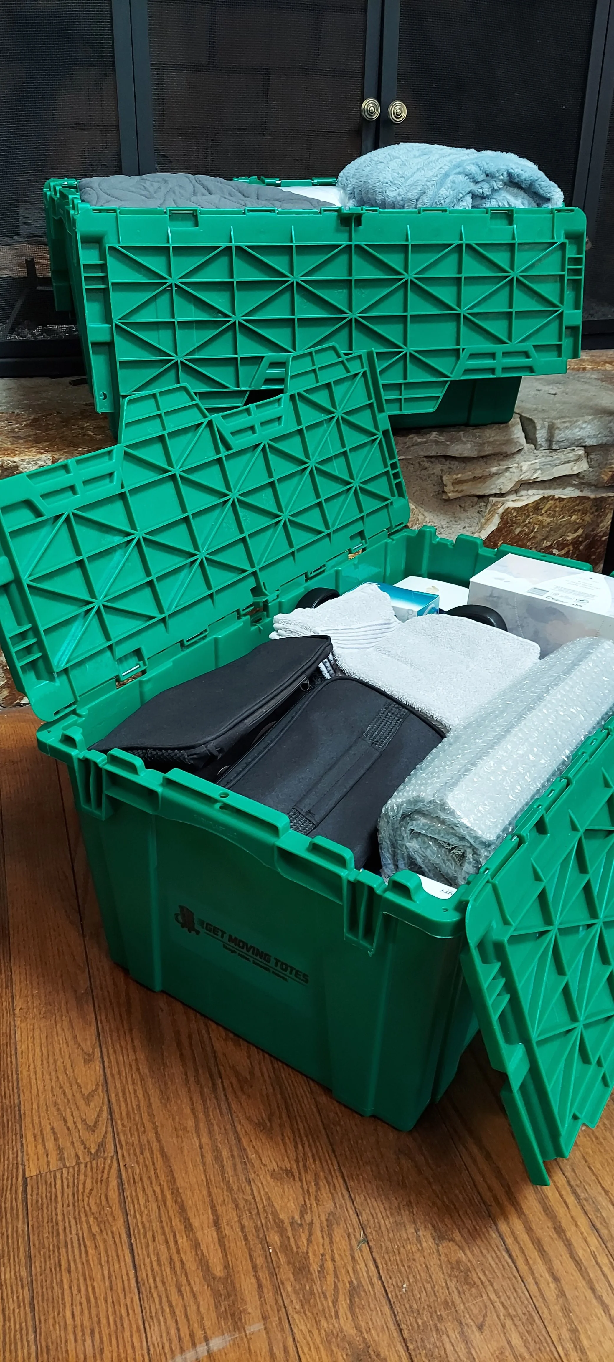 Open green plastic moving tote container filled with household items including a black case, white cloths, a bubble wrap roll, and a box, placed on a wooden floor in front of a stone hearth and black fireplace doors.