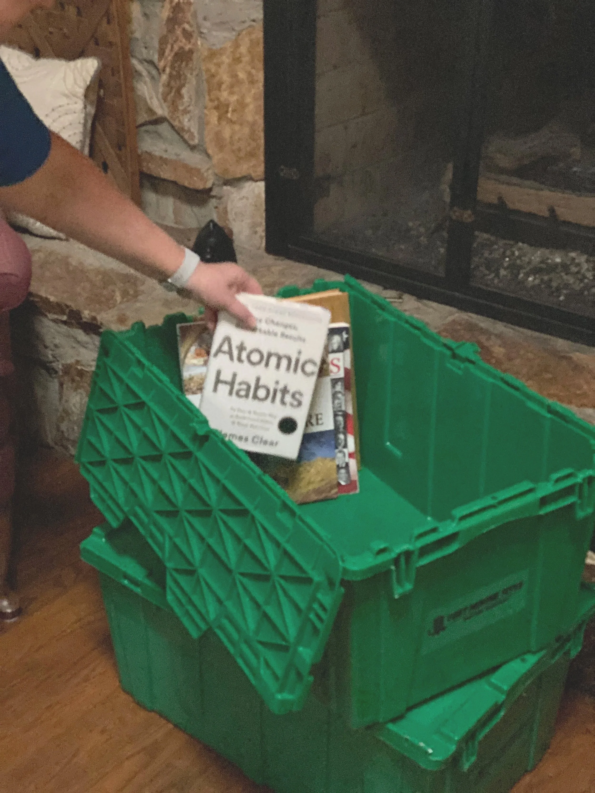 A green plastic crate filled with books about environmental issues, including one titled 'Atomic Habits', next to a fireplace with stone and brick around it.