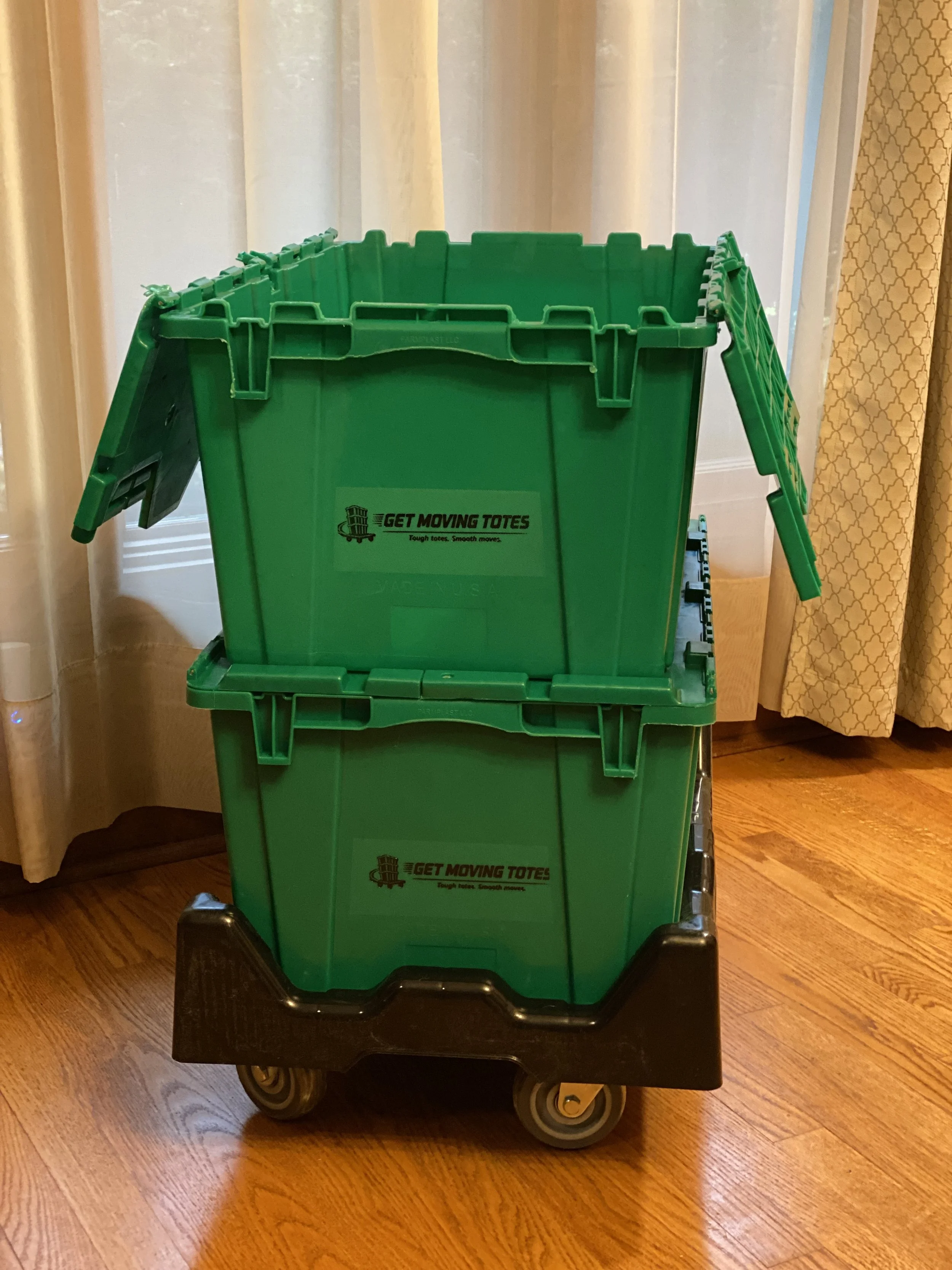A green plastic storage tote on a black wheeled platform with a handle, placed on a wooden floor indoors. The tote has a label that reads 'GET MOVING TOTES' with the tagline 'Tough totes. Smooth moves.'