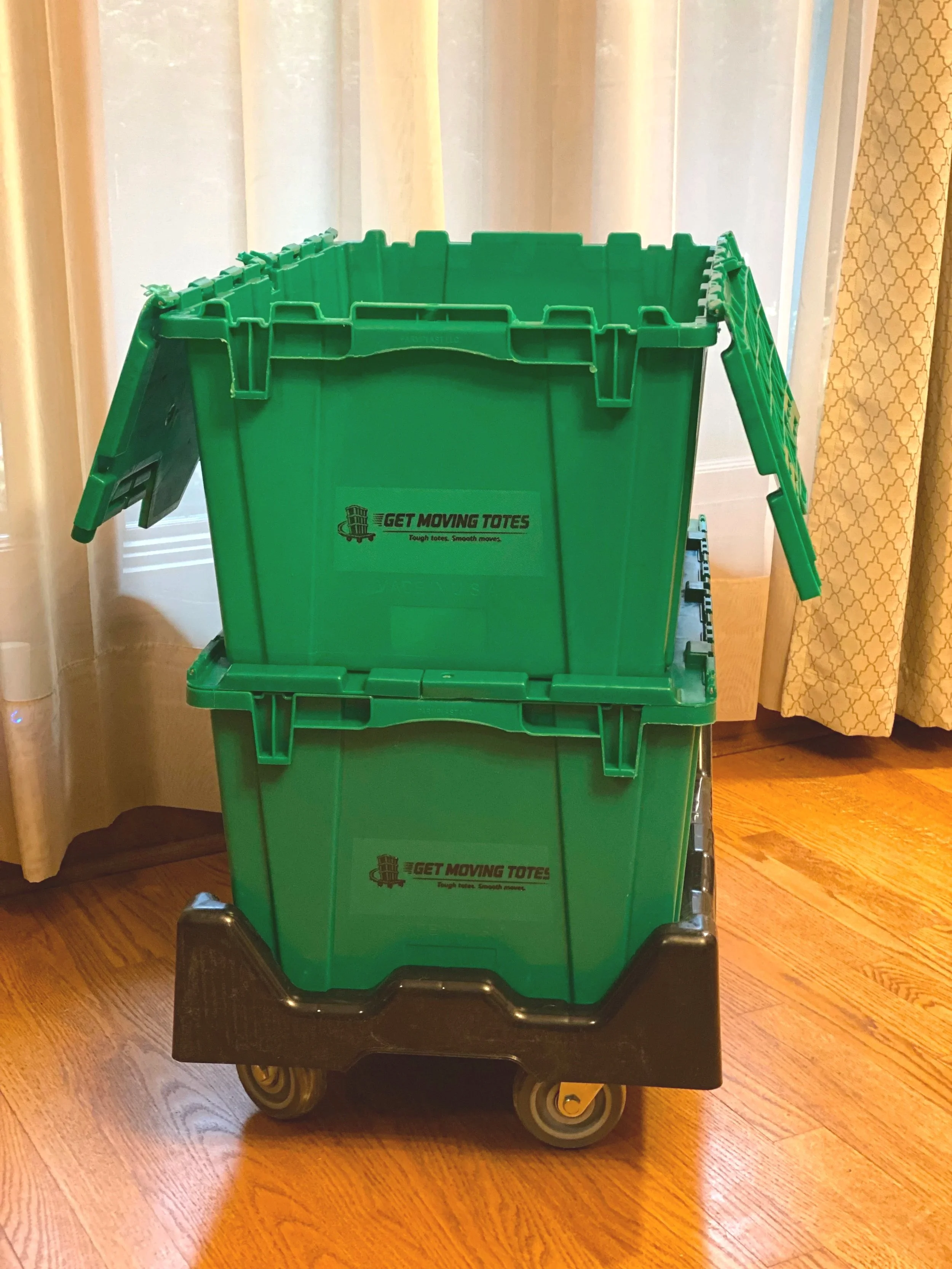 Green moving tote box on a black rolling dolly with wood flooring and curtains in the background.