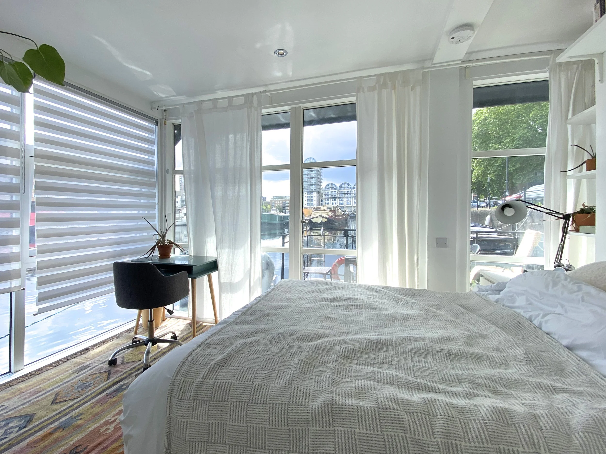 A bedroom with large windows showing a river and buildings outside. The bed has a textured blanket, and there is a black chair at a small green desk with a potted plant. White curtains and blinds cover the windows. The room has a colorful rug on a wooden floor.
