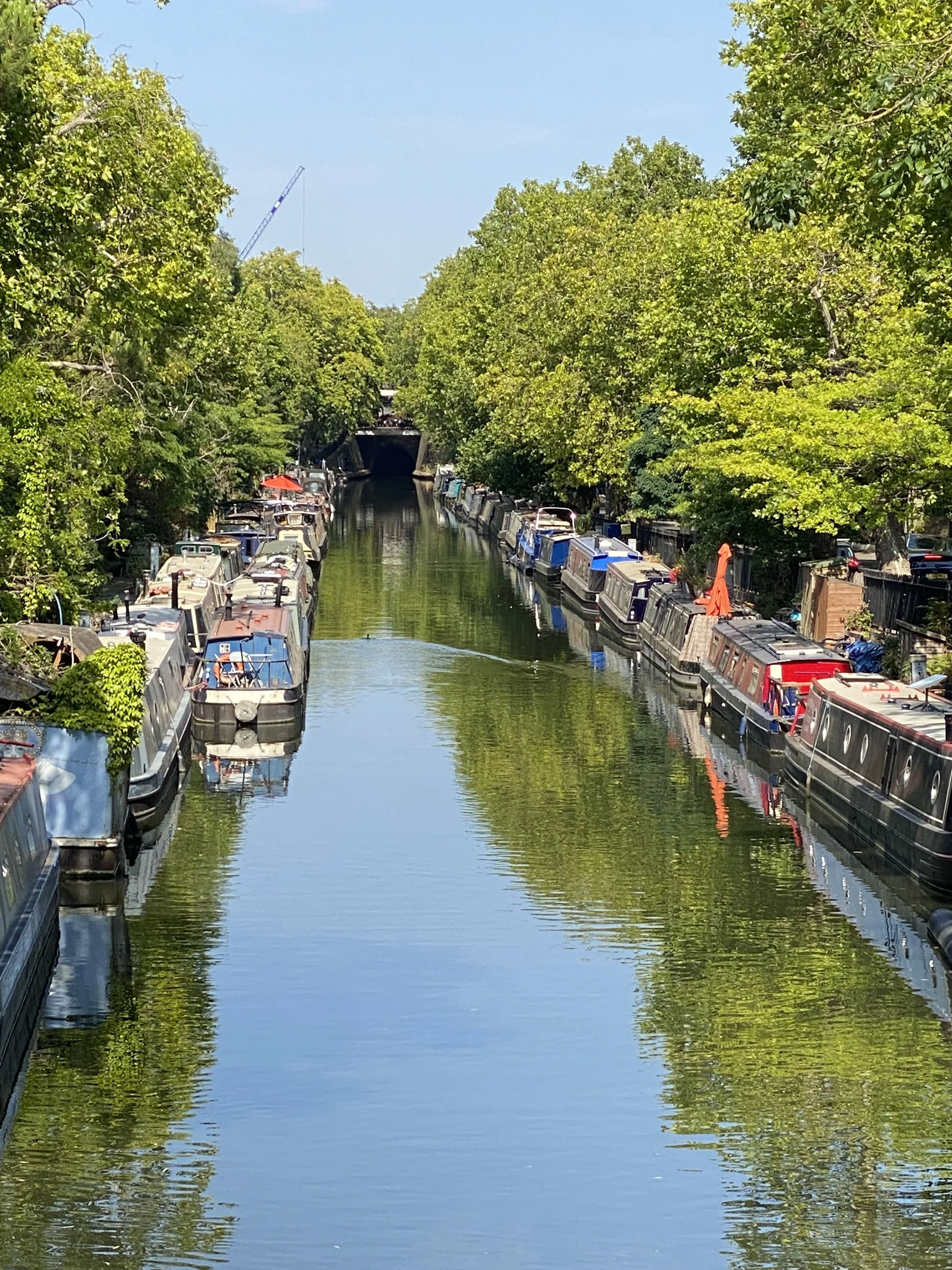 A canal lined with trees and moored boats on both sides, with a tunnel in the background and a crane visible above the trees.