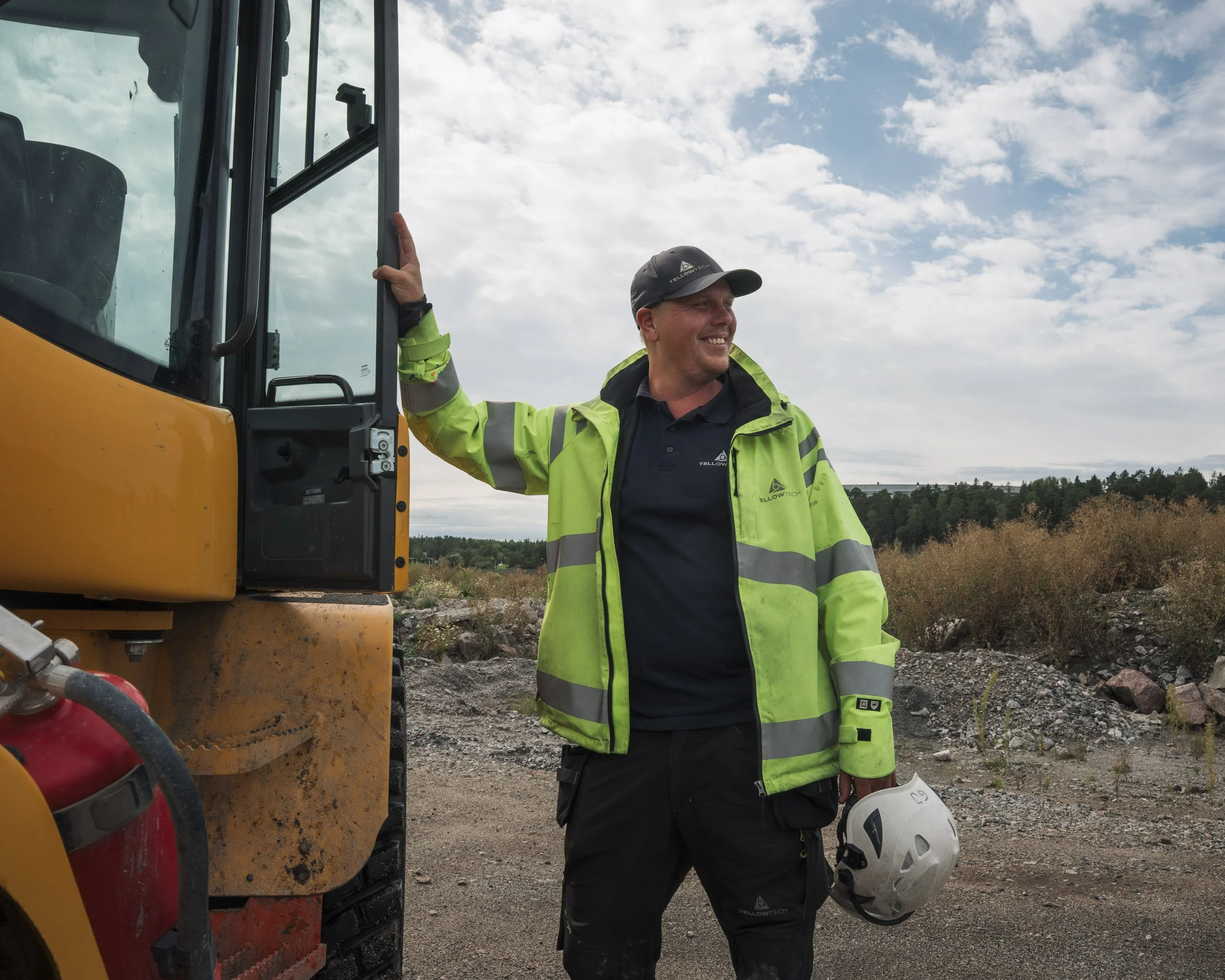 A smiling man wearing a high-visibility jacket and cap standing next to construction equipment outdoors on a partly cloudy day.