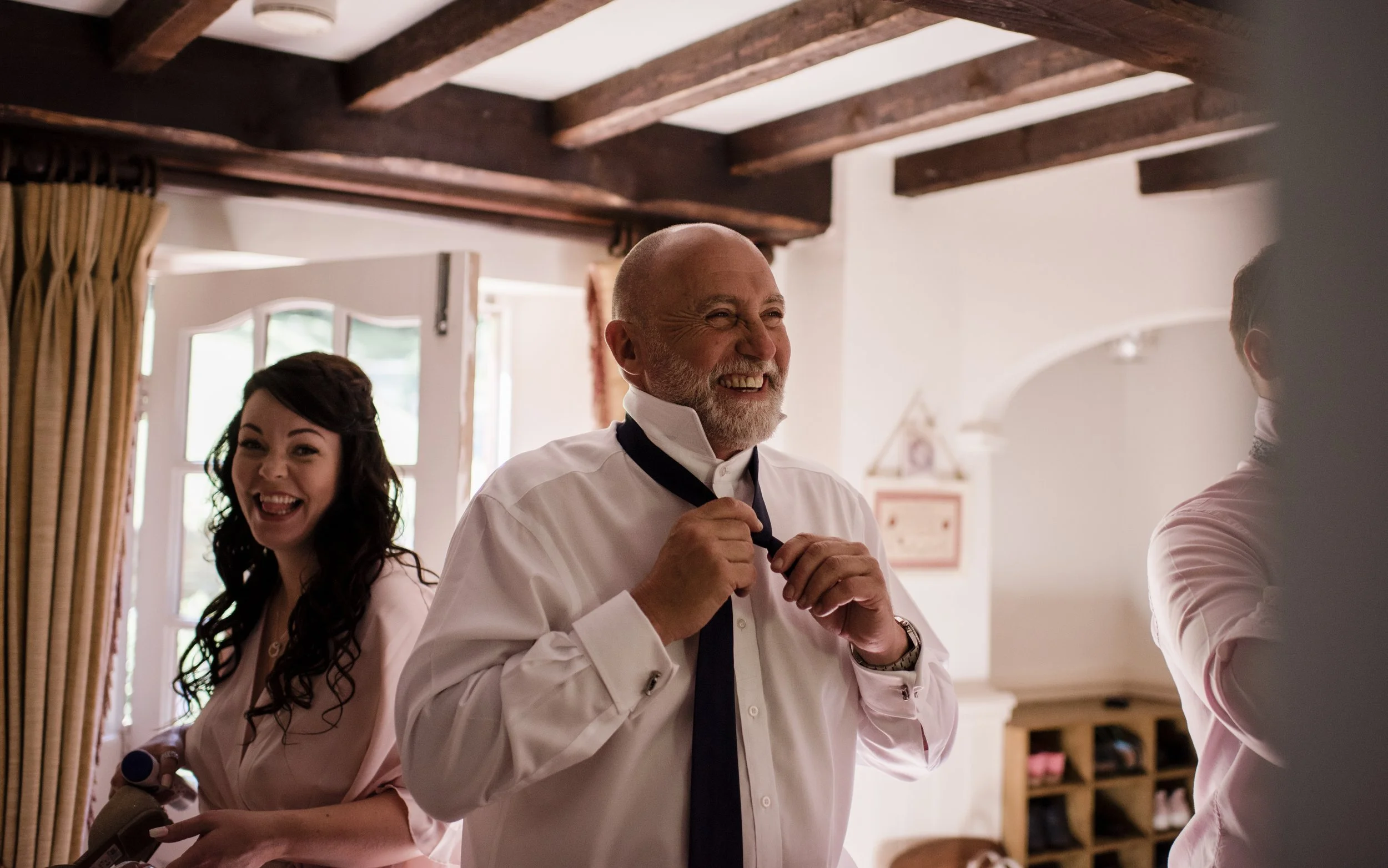 A joyful man with a white beard and smiling woman with dark hair, both in formal attire, standing in a warmly lit room with wooden beams on the ceiling and a window with curtains.