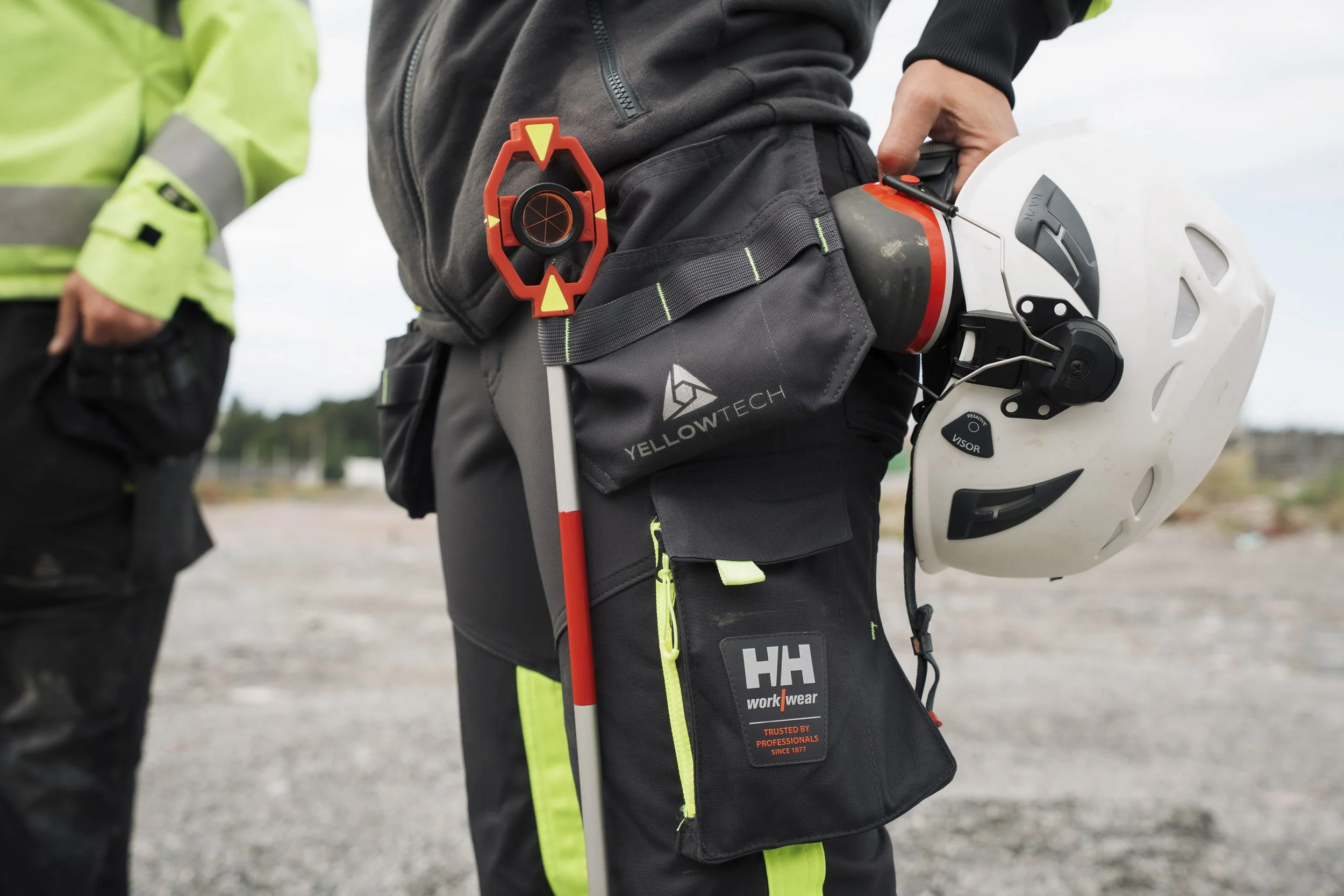 Close-up of a person holding a white safety helmet in their right hand, with a black work pouch attached to their black pants, carrying safety gear including a bright orange safety device and a black and red tool. The person is dressed in black and s