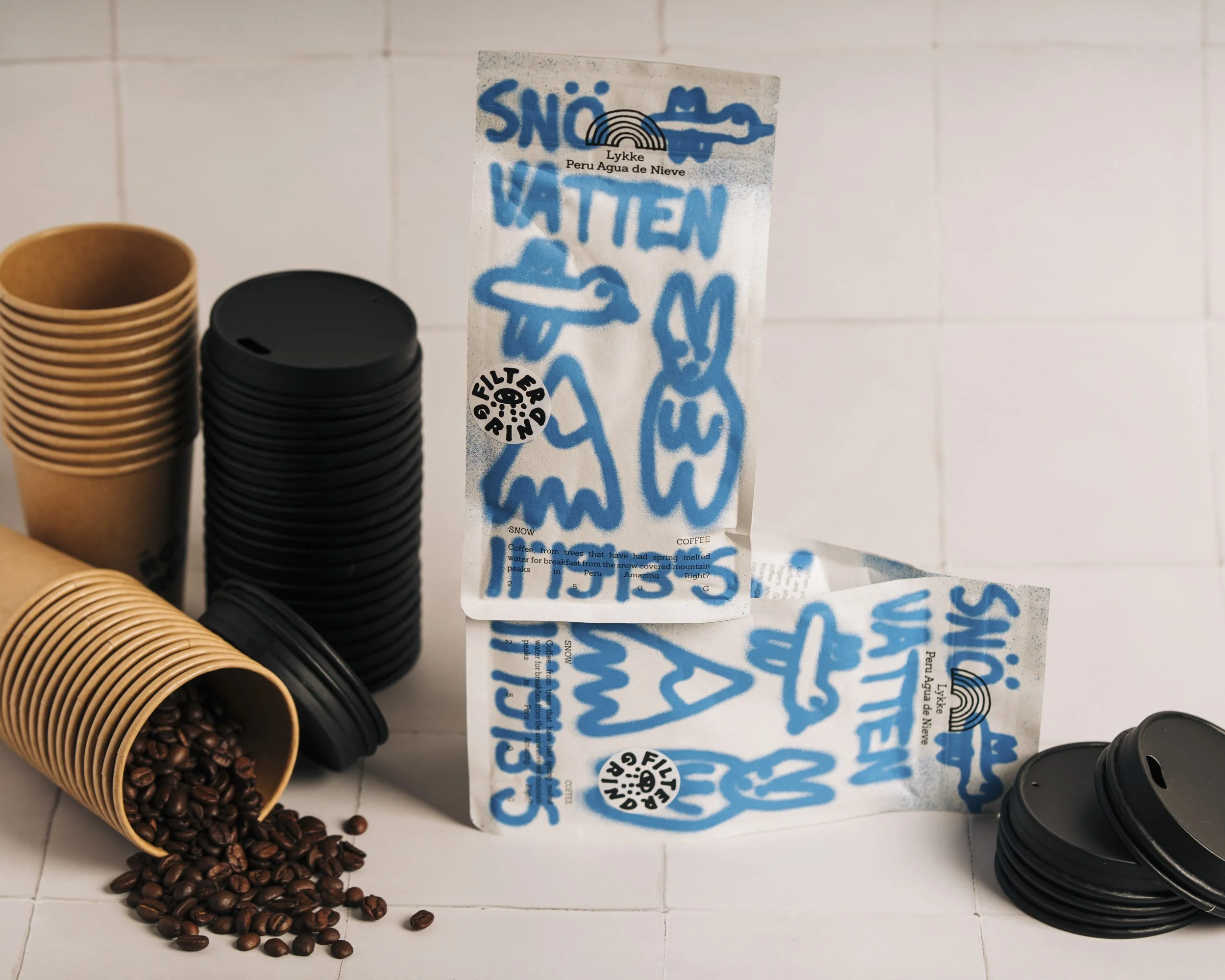 Disposable coffee cup on its side with coffee beans spilled out, beside another stacked disposable cup and a paper bag with blue lettering and logo, placed on a white tiled surface.