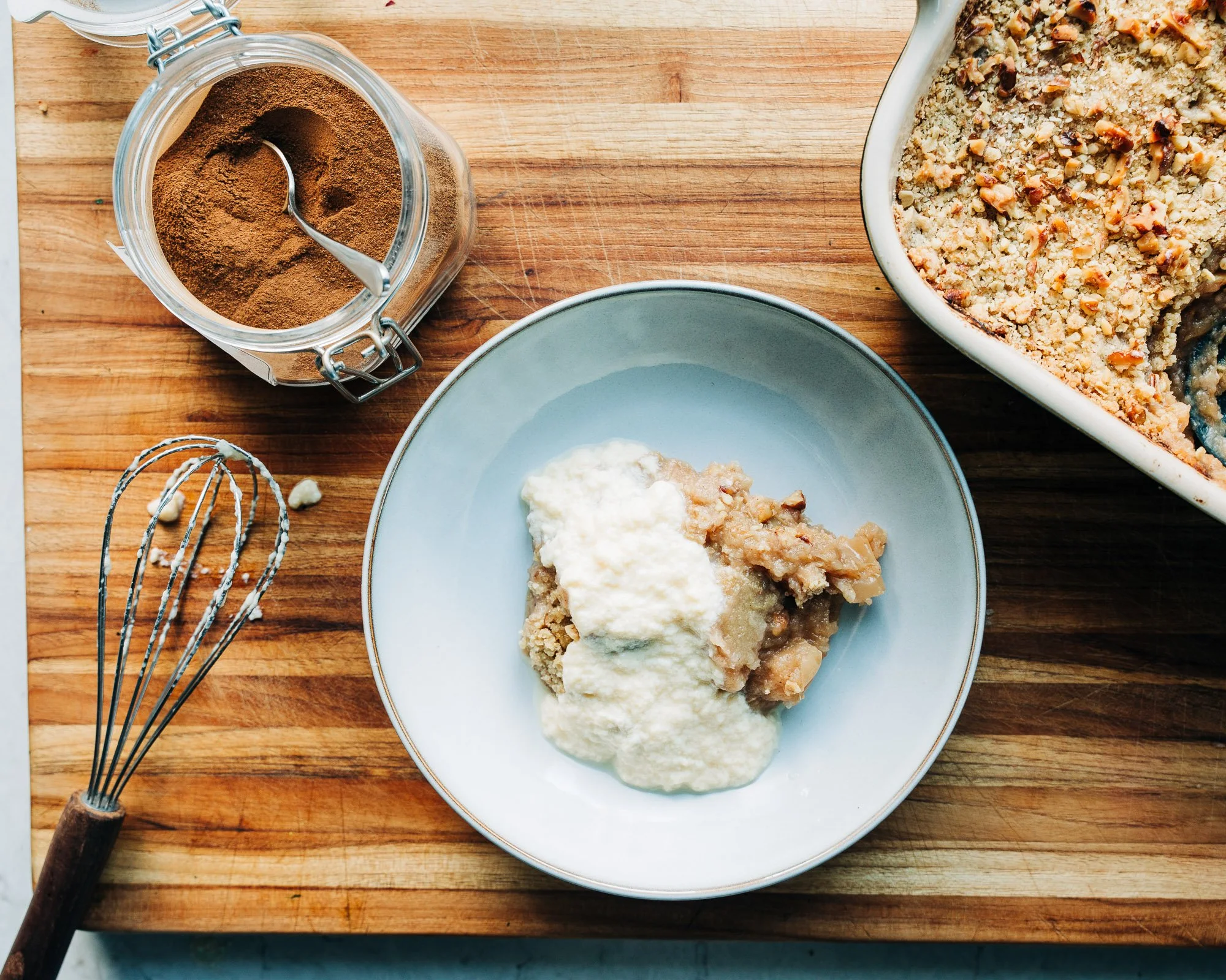 A wooden cutting board with a partially baked fruit crumble in a white dish, a bowl of vanilla ice cream, a jar of ground cinnamon, and baking utensils including a whisk and a hand-held electric mixer.