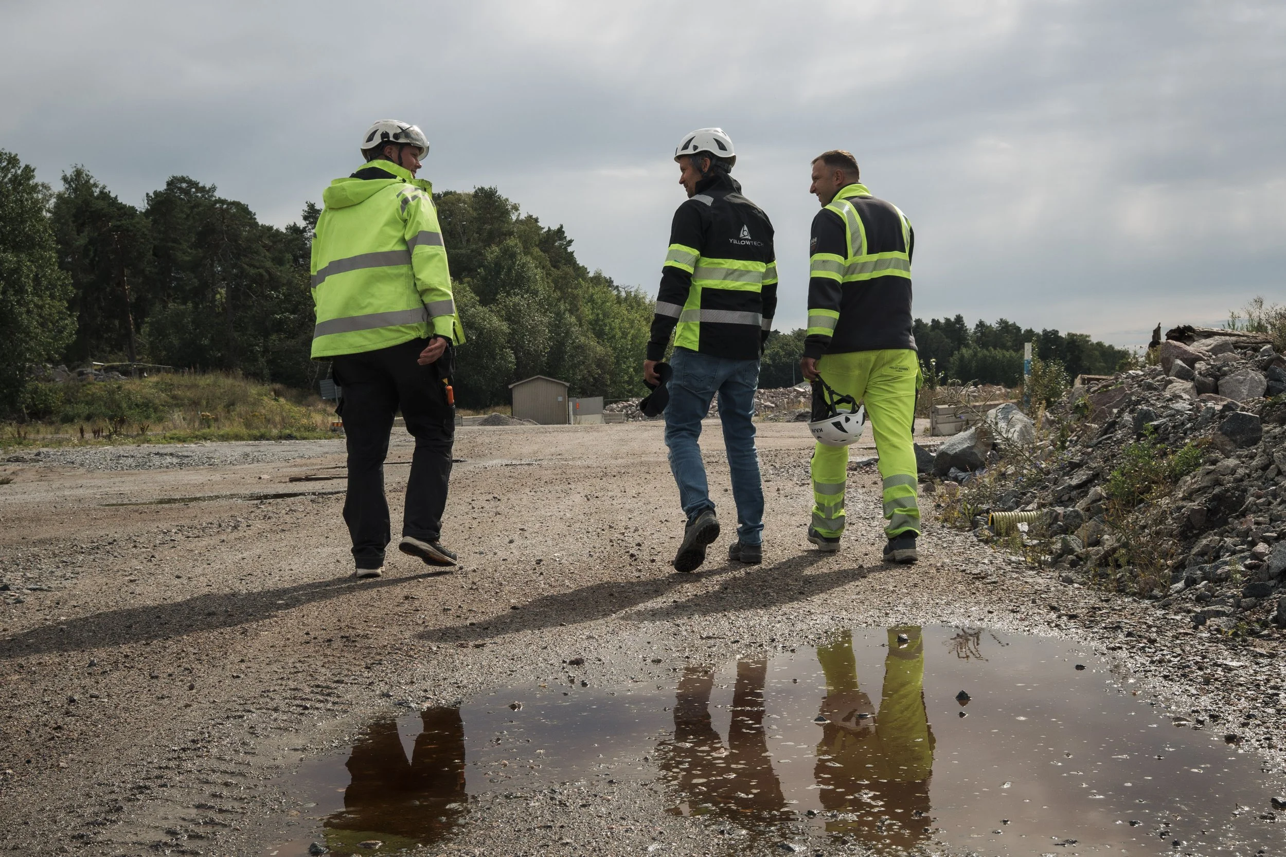 Three construction workers wearing helmets and high-visibility jackets standing on a dirt road near a small pool of muddy water, with rocks and vegetation in the background.