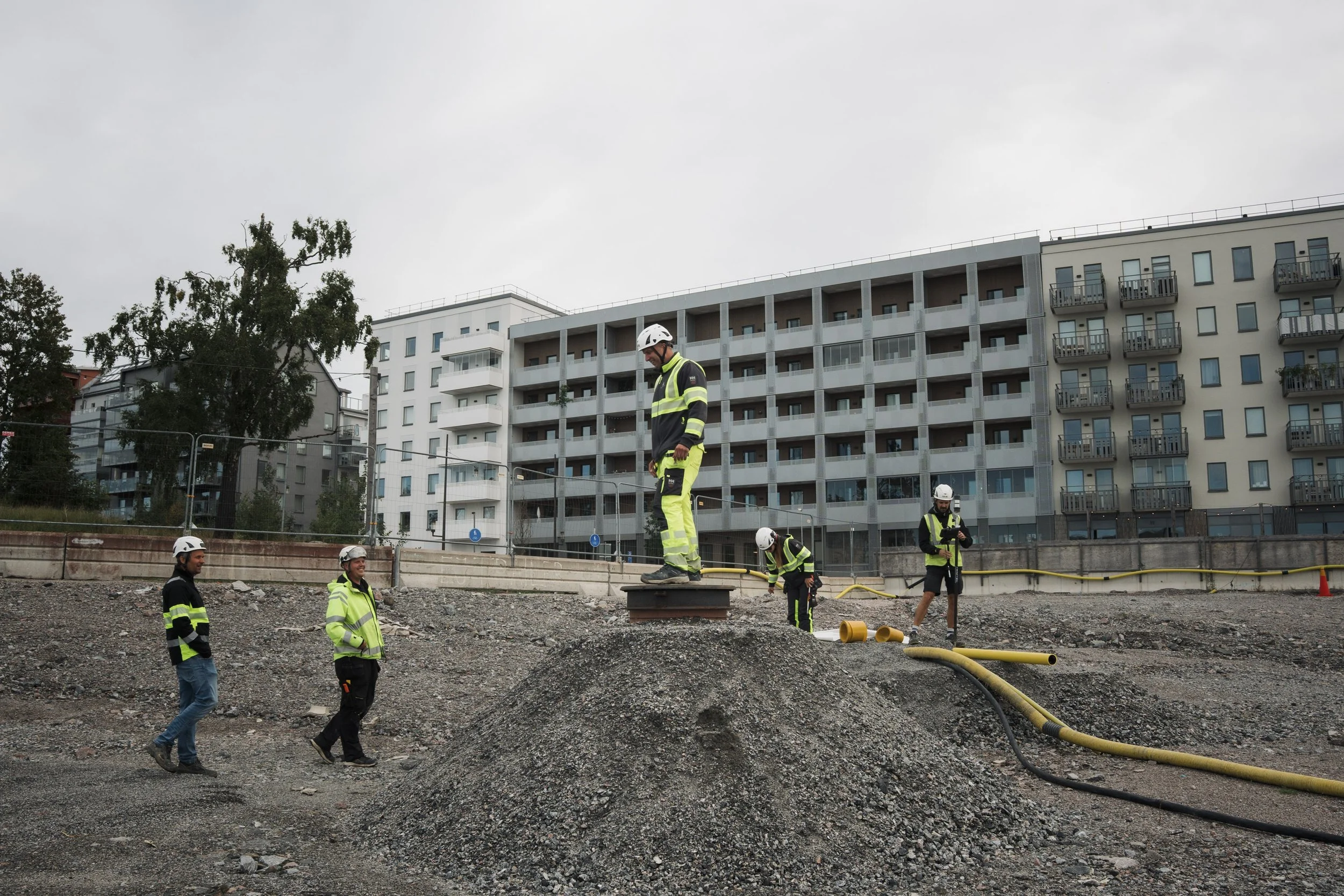 Workers in safety gear working on a construction site in front of modern residential buildings under cloudy skies.