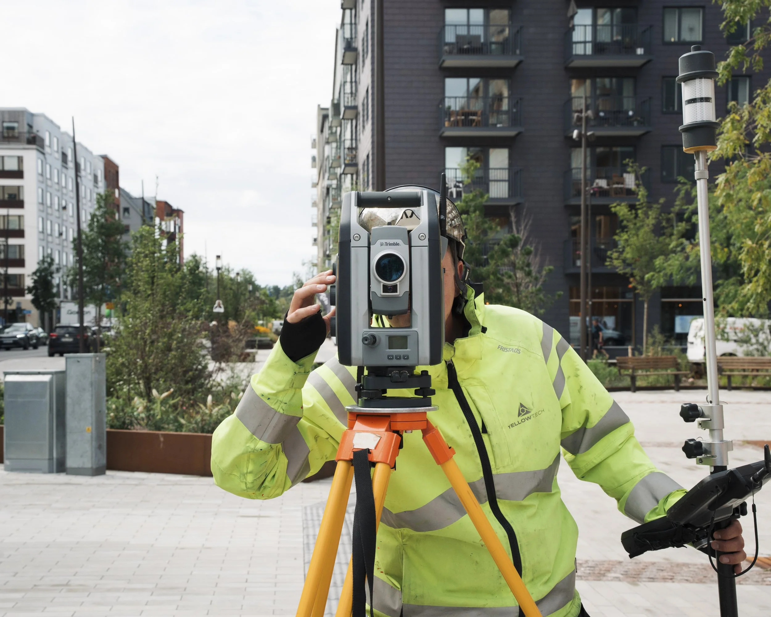 A construction worker wearing a high-visibility yellow jacket and helmet using a total station survey instrument on a city street with modern apartment buildings in the background.