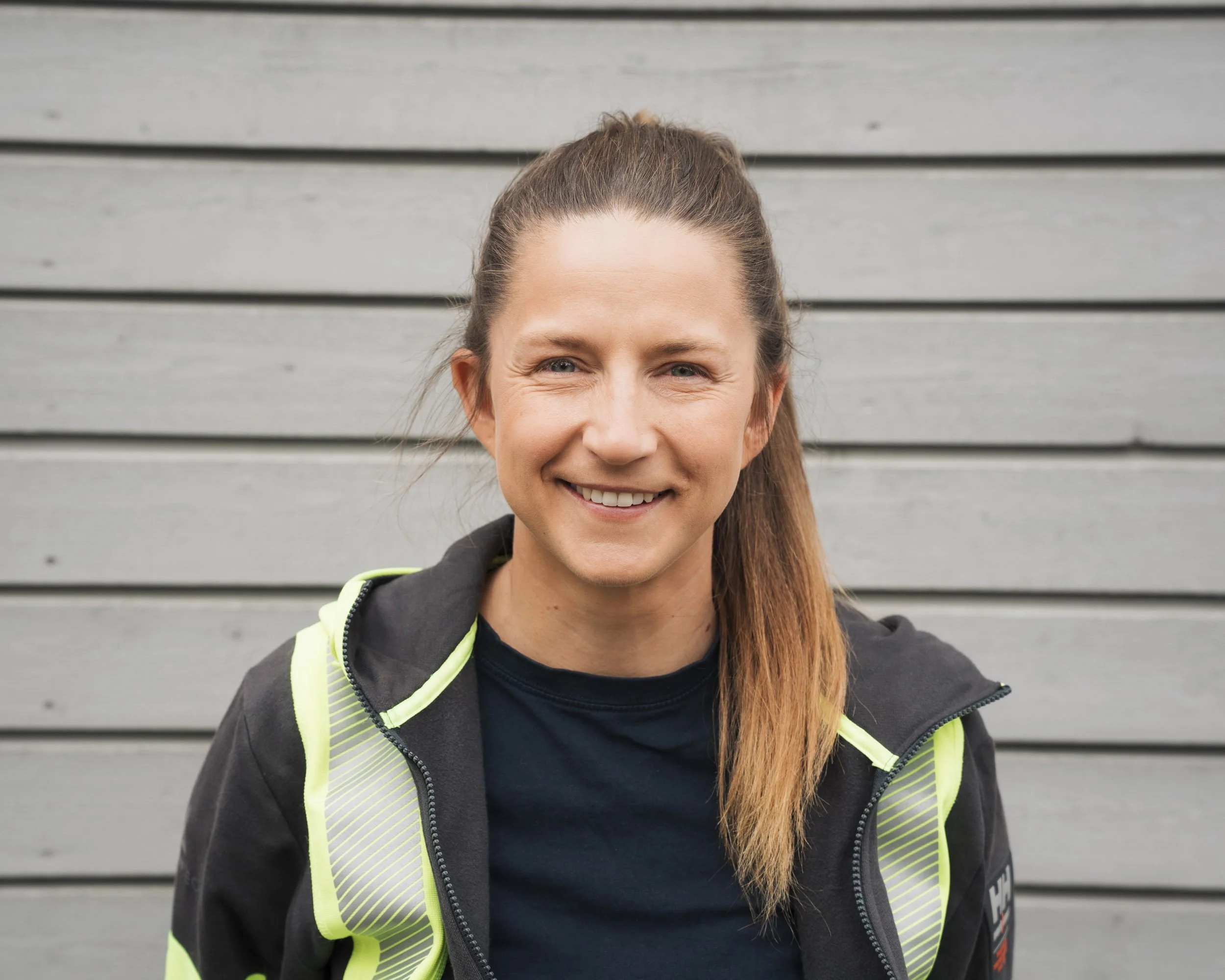 Close-up of a smiling woman with light brown hair tied back, wearing a black shirt and a neon yellow and black jacket, standing in front of a gray horizontal wooden plank background.