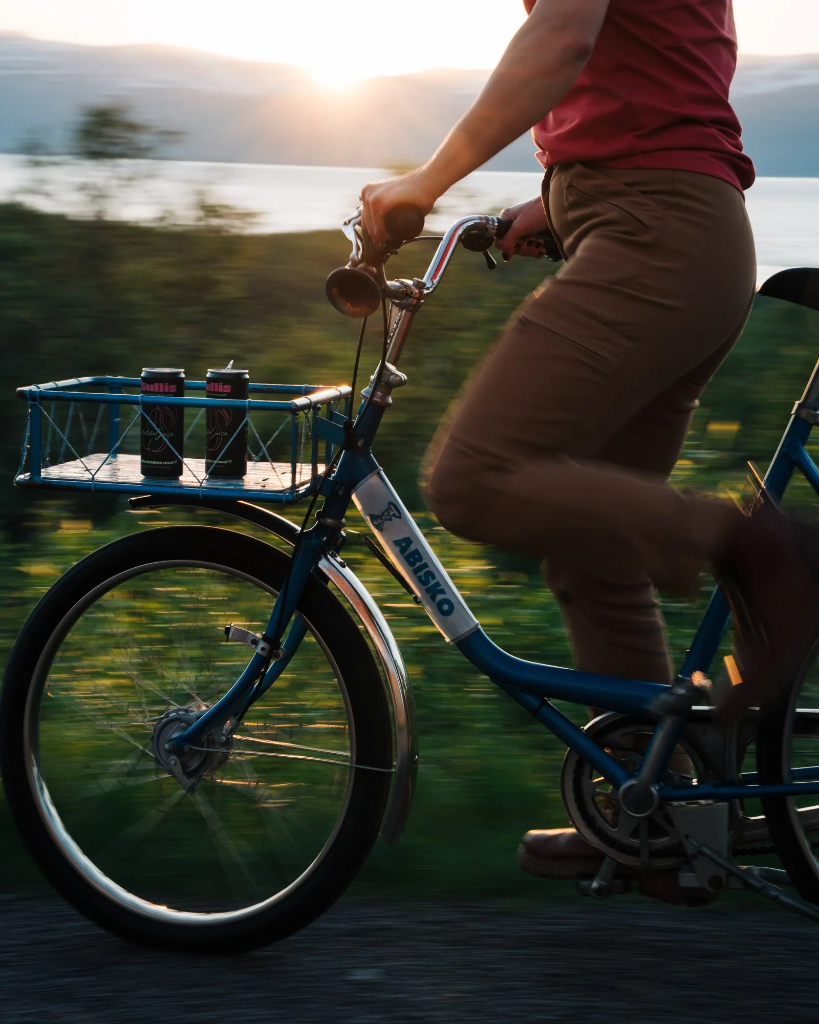 Close-up of a person riding a bicycle outdoors at sunset with cans of beverage in a front rack.