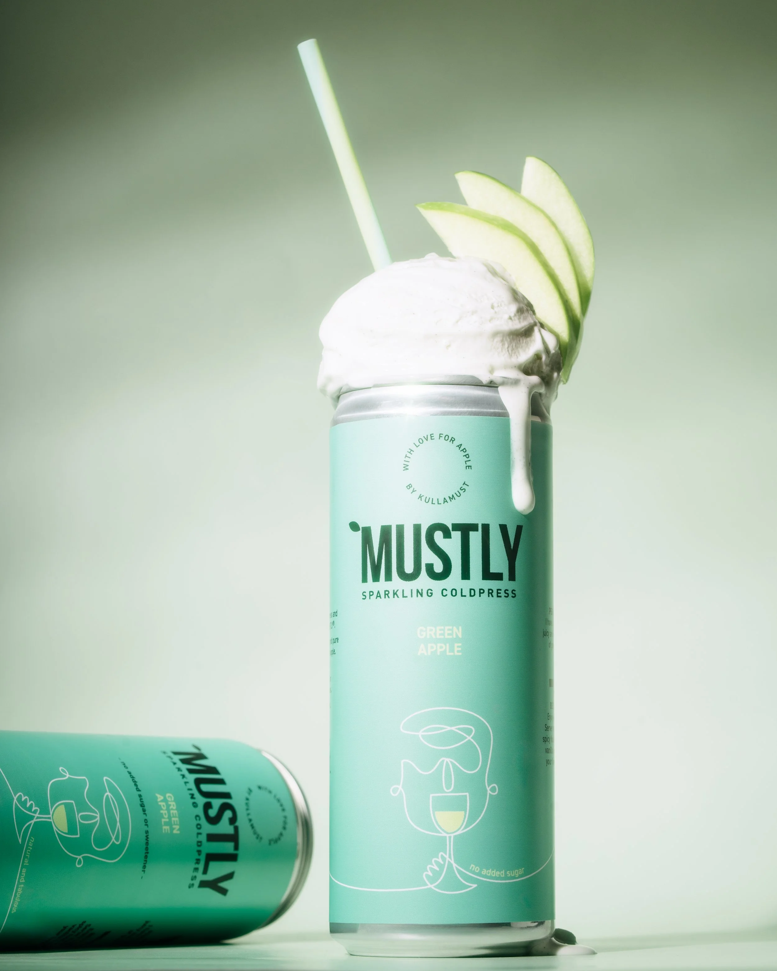 A can of Mustly sparkling coldpress green apple beverage with foam and slices of green apple on the rim, against a light green background.