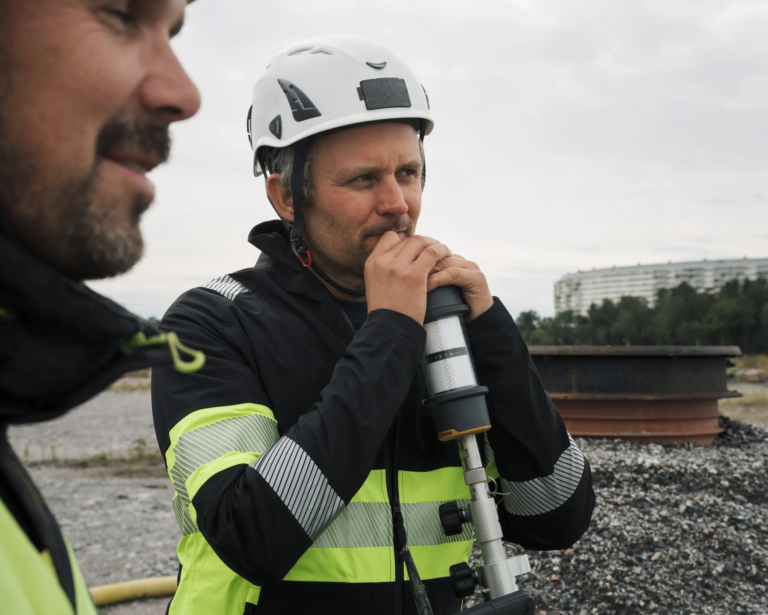 Two men wearing high-visibility jackets and construction helmets, with one holding a surveying instrument outdoors against a cloudy sky and distant buildings.