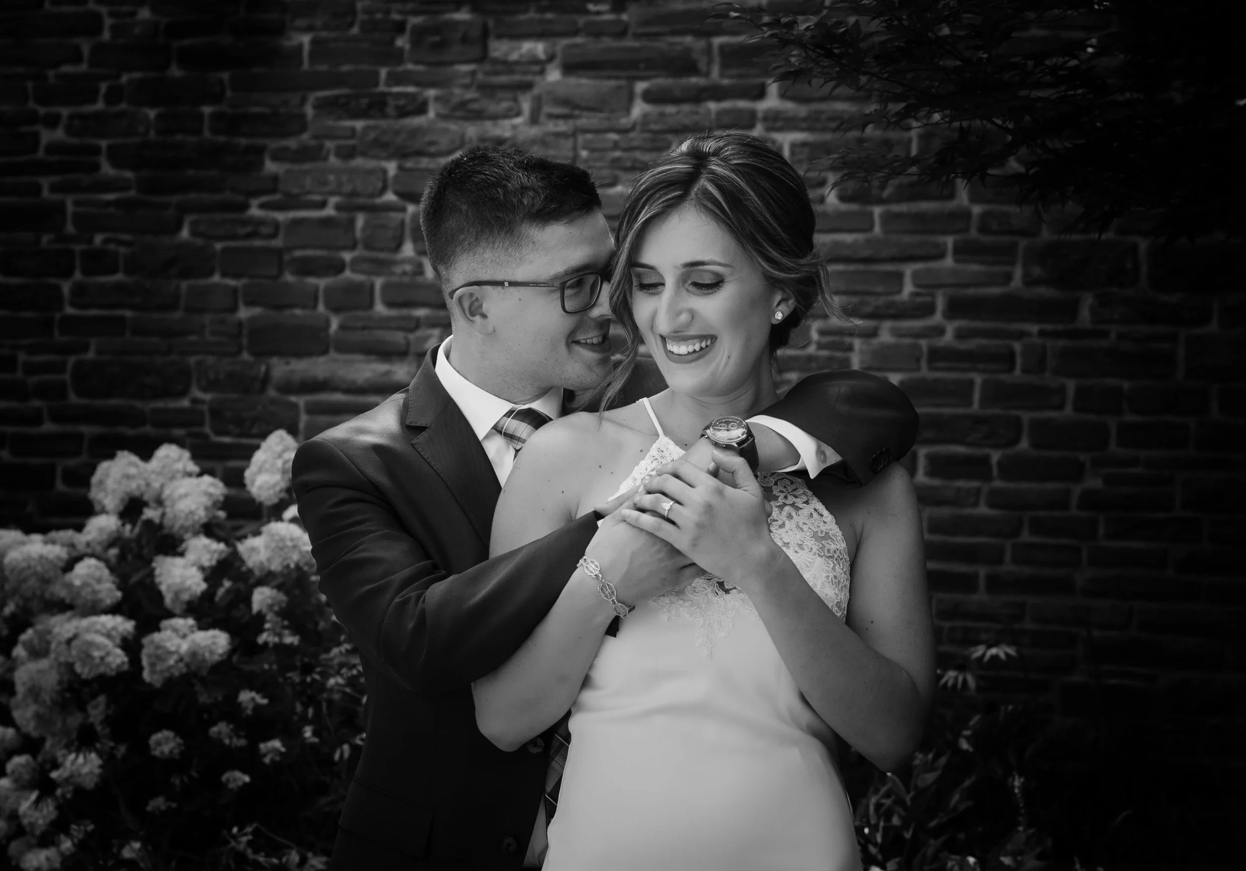 A black and white photo of a smiling couple, with the man embracing the woman from behind, holding her hand, in front of a brick wall. The woman is wearing a strapless dress, and the man is in a suit with glasses.