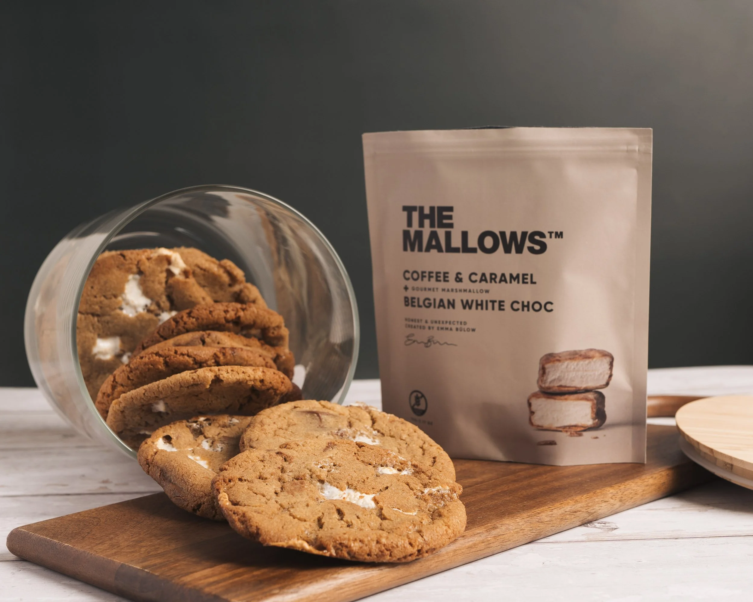Cookies with marshmallow filling on a wooden cutting board, a bag of coffee and caramel flavored marshmallow, and a partial view of a glass container with more cookies.